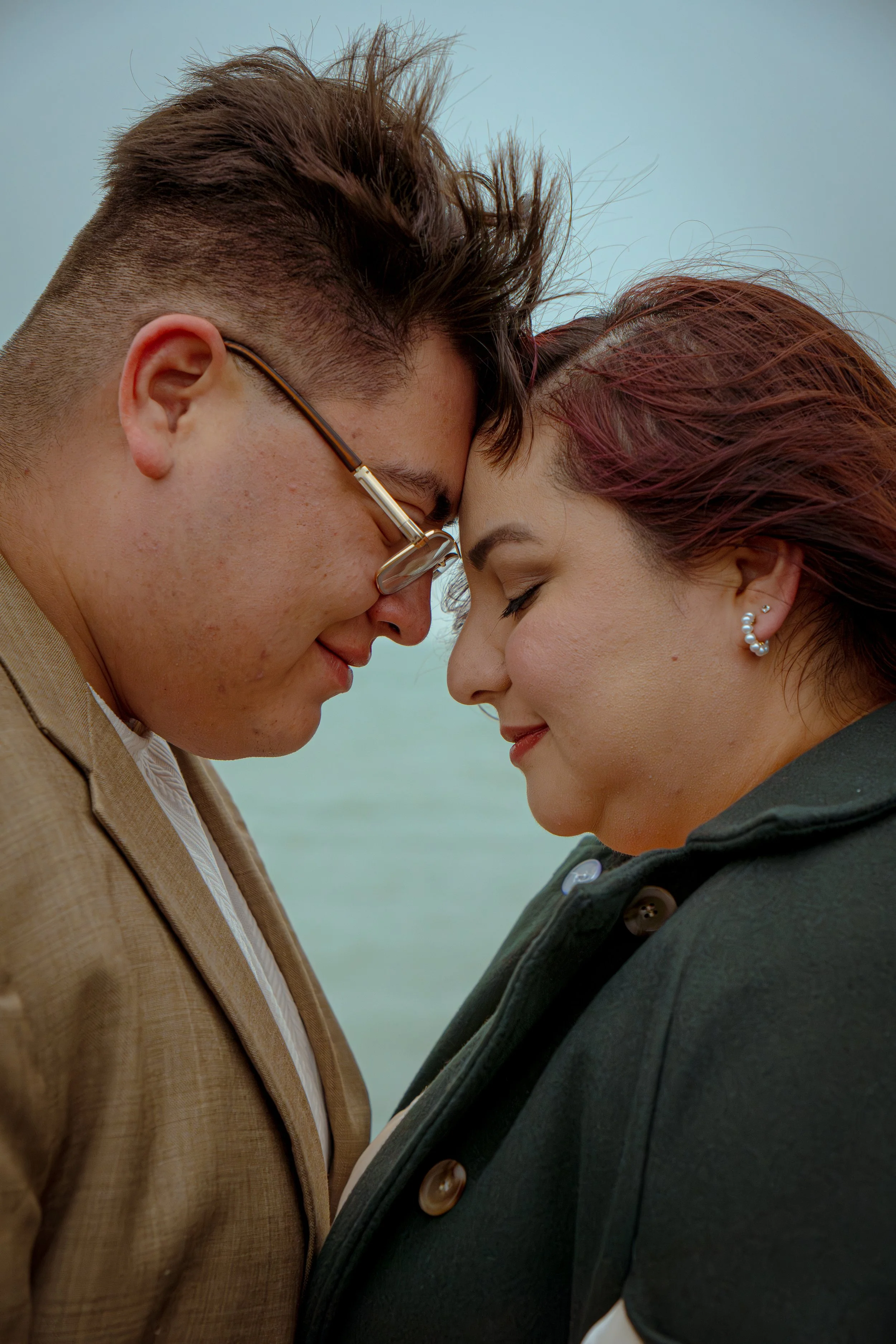 A close-up of two people touching foreheads with eyes closed, smiling and sharing an intimate moment. The person on the left is wearing glasses and a tan jacket, and has short, styled hair. The person on the right has reddish hair and is wearing a da