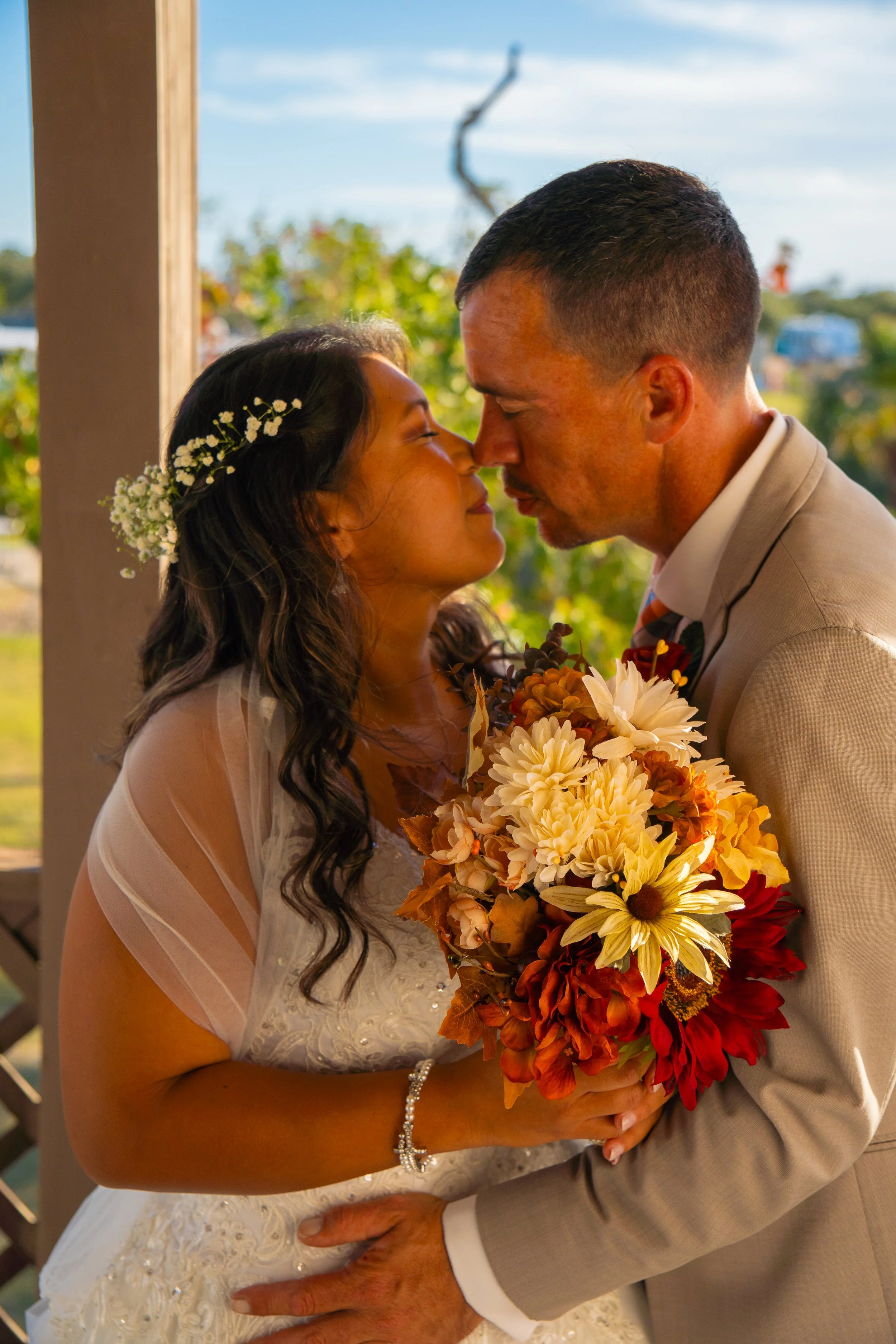 A bride and groom sharing a tender moment with their noses touching, holding a vibrant bouquet of red, orange, white, and yellow flowers, during their wedding.