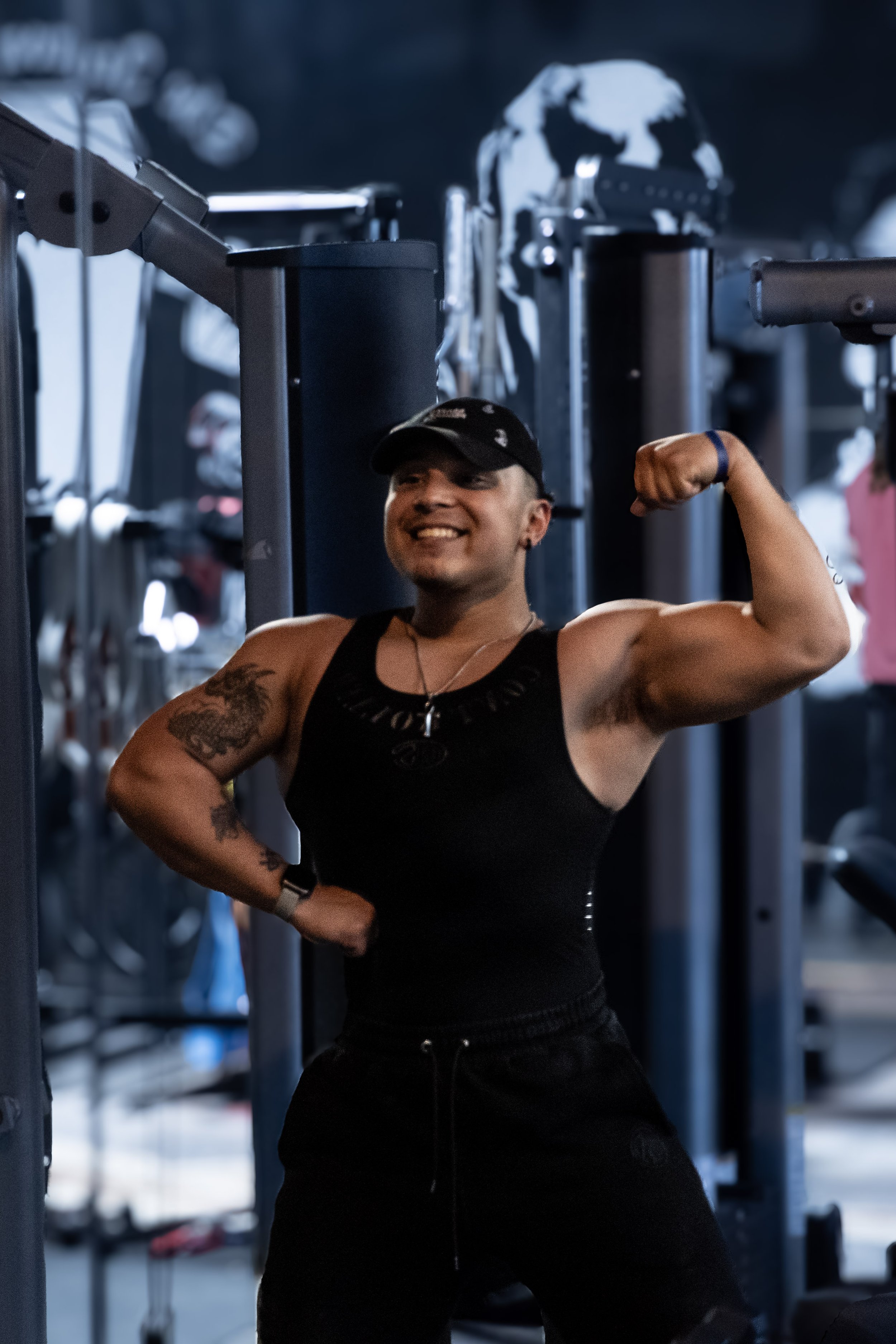 A woman with tattoos on her arms, wearing a black tank top and cap, smiling and flexing her biceps in a gym.