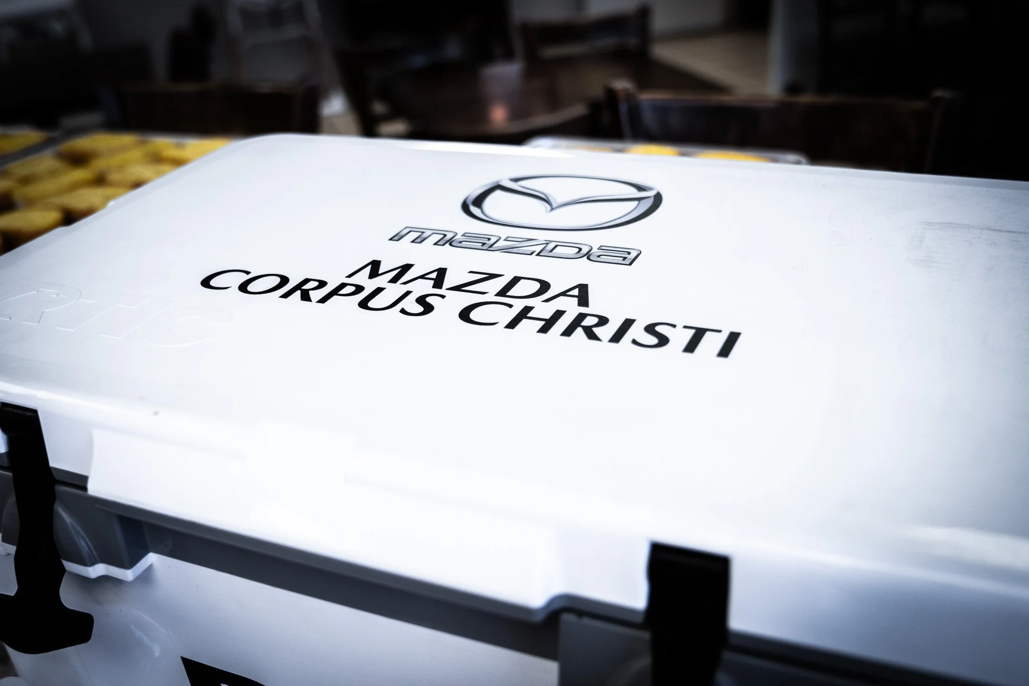 Close-up of a white box with the Mazda logo and the text "Mazda Mazda Corpus Christi" printed on it, placed on a table with some food items in the background.