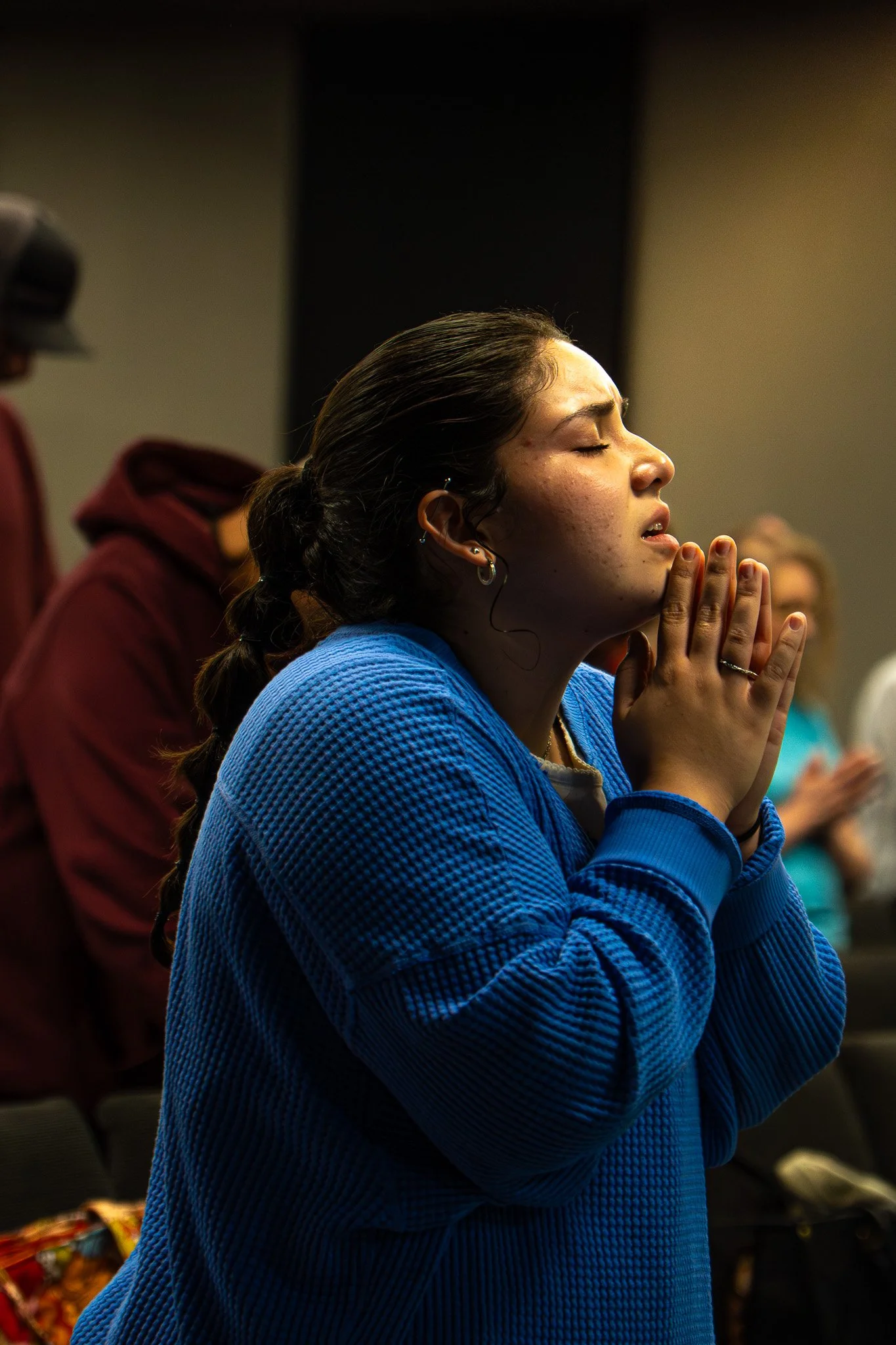 A young woman with dark hair pulled back, wearing a blue sweater, praying with her eyes closed and hands clasped together.