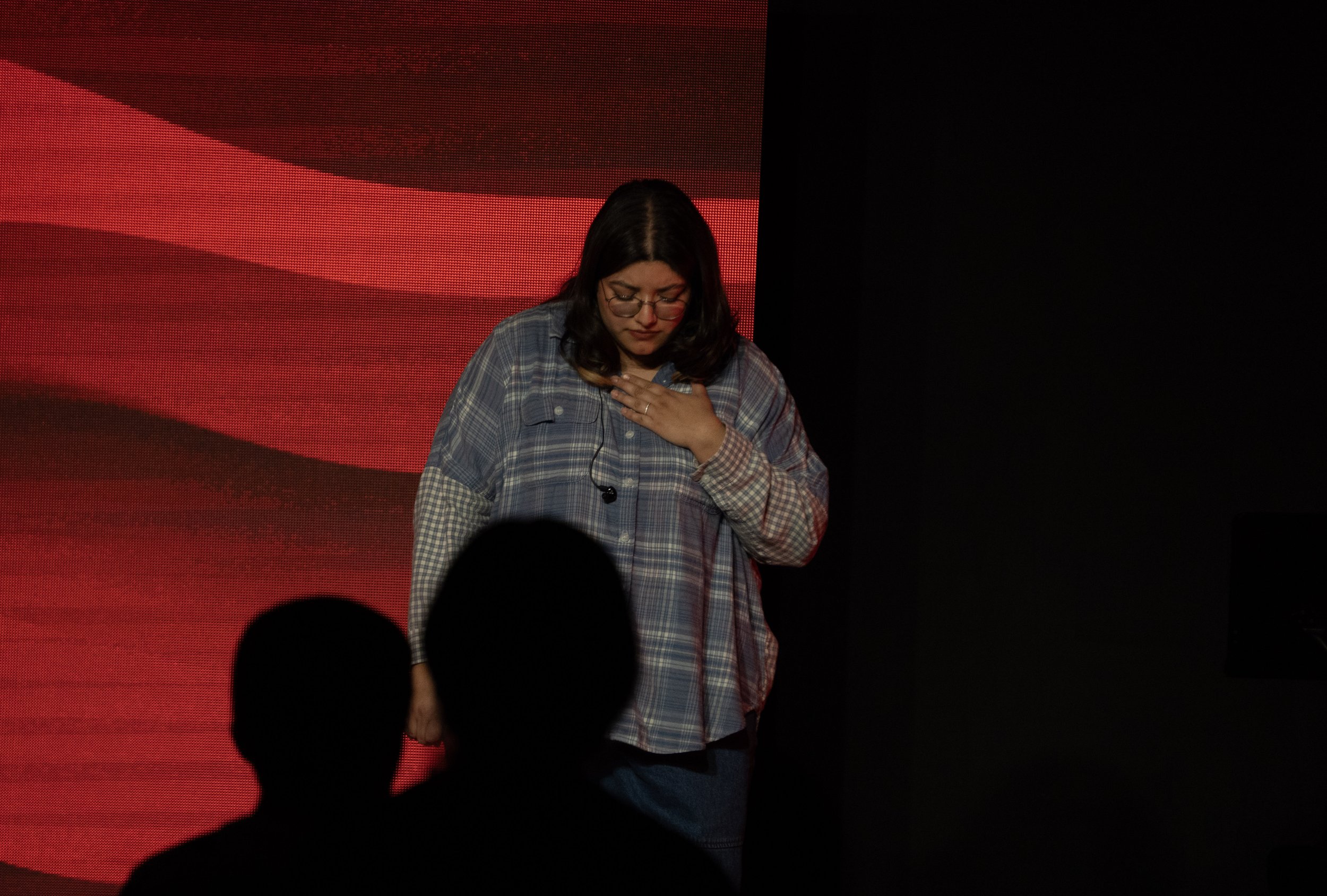 A woman wearing glasses and a plaid shirt standing on stage with a hand on her chest, with an American flag background behind her.