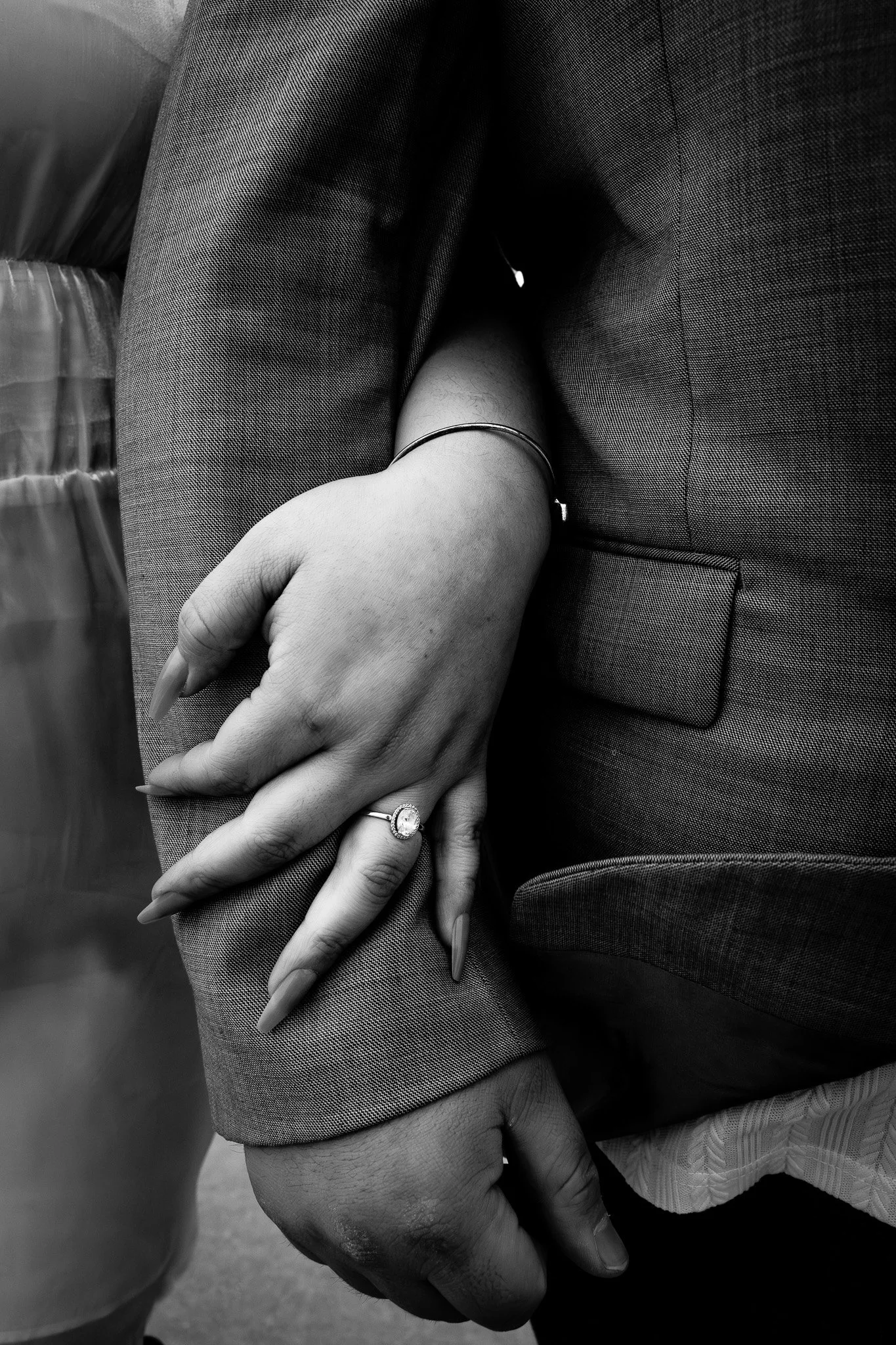 Close-up of two hands intertwined, one with an engagement ring and jewelry, resting against a person's sleeve in a professional suit.
