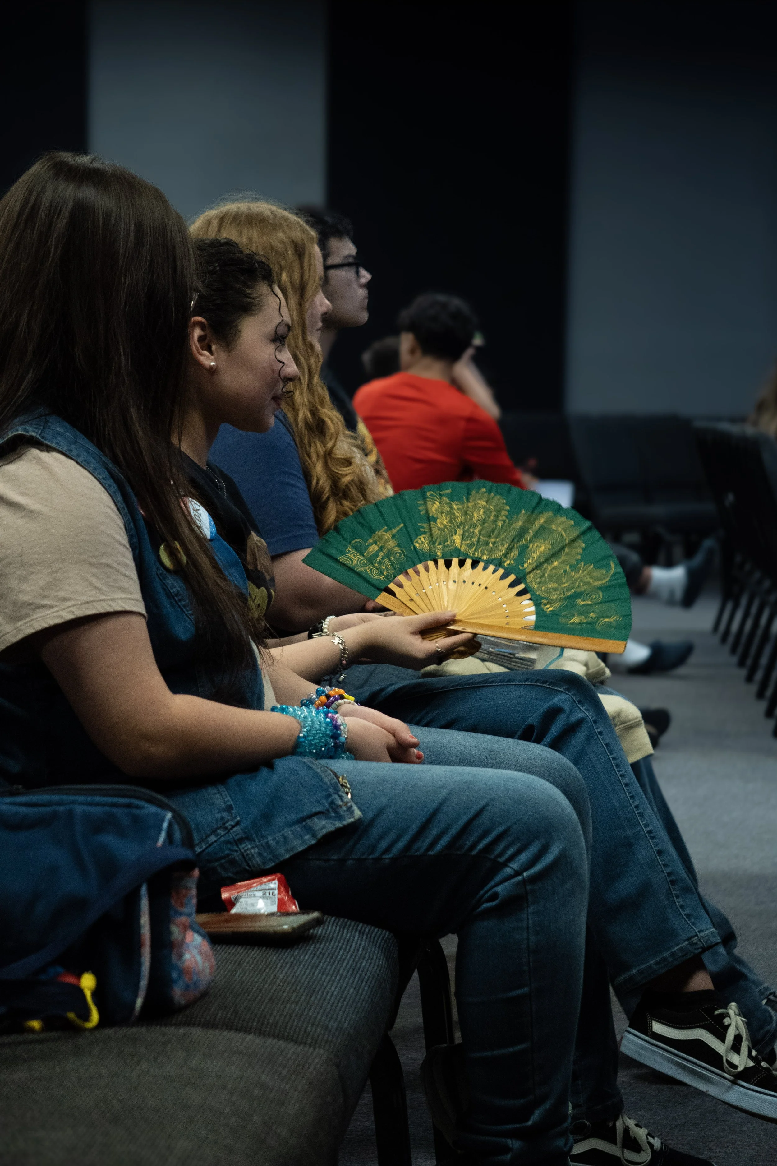 A group of teenagers or young adults sitting in a row during a presentation or event, with one holding a green decorative fan.