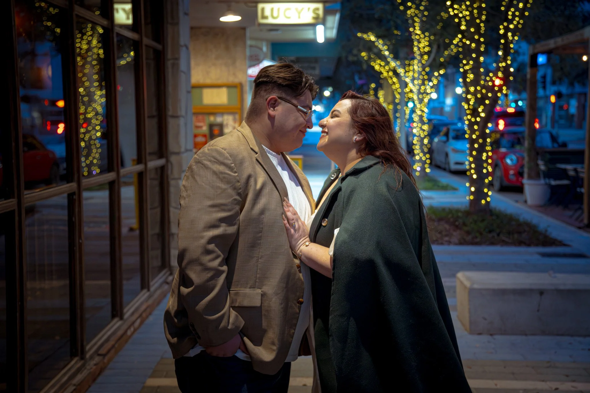A couple standing close and smiling at each other on a city street at night, decorated with string lights on trees, with parked cars and a restaurant with a sign that reads 'LUCY'S' in the background.