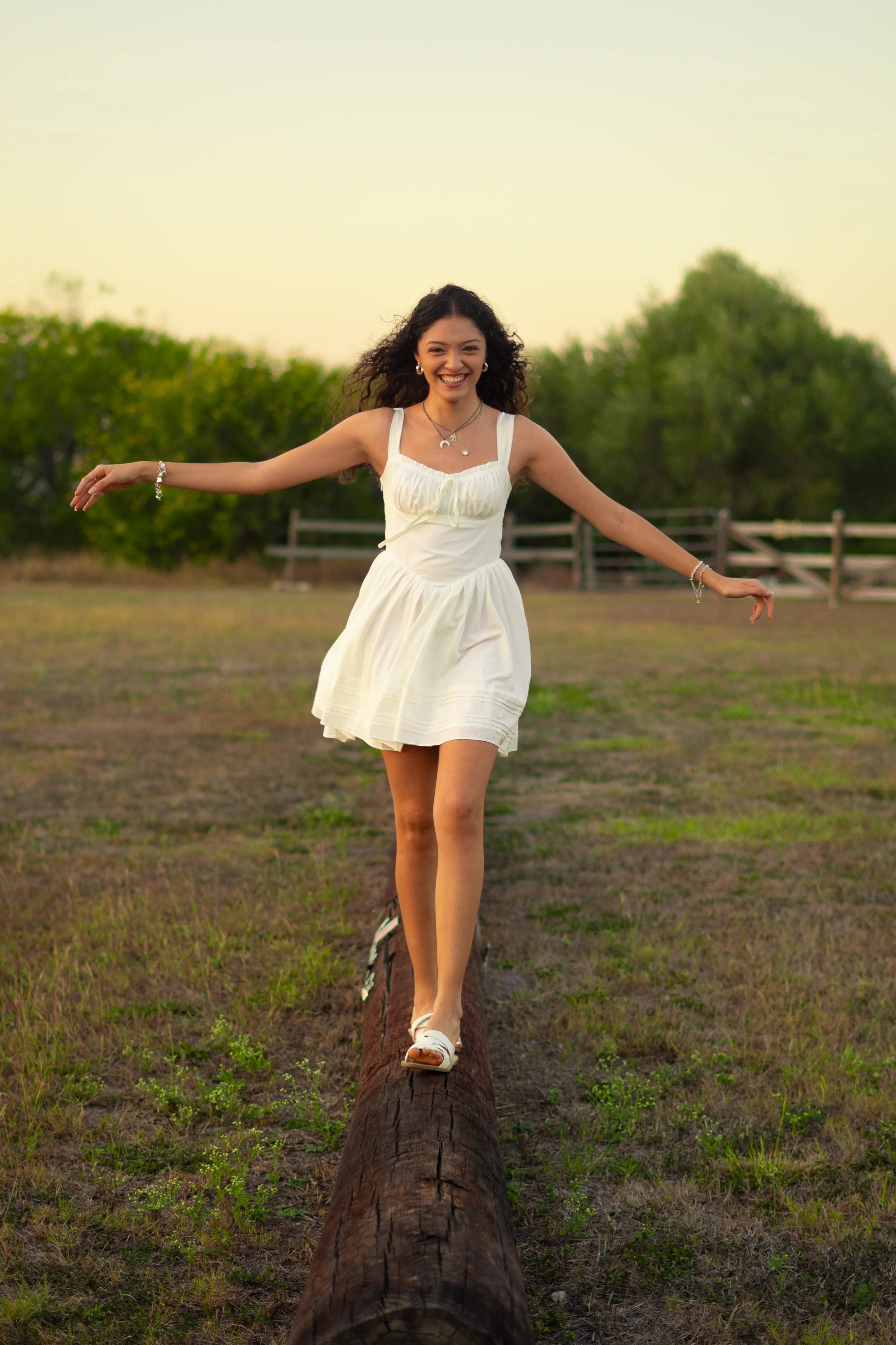 A young woman with curly hair smiling and extending her arms outward while walking on a log in an outdoor grassy area with trees and a fence in the background during late afternoon or early evening.