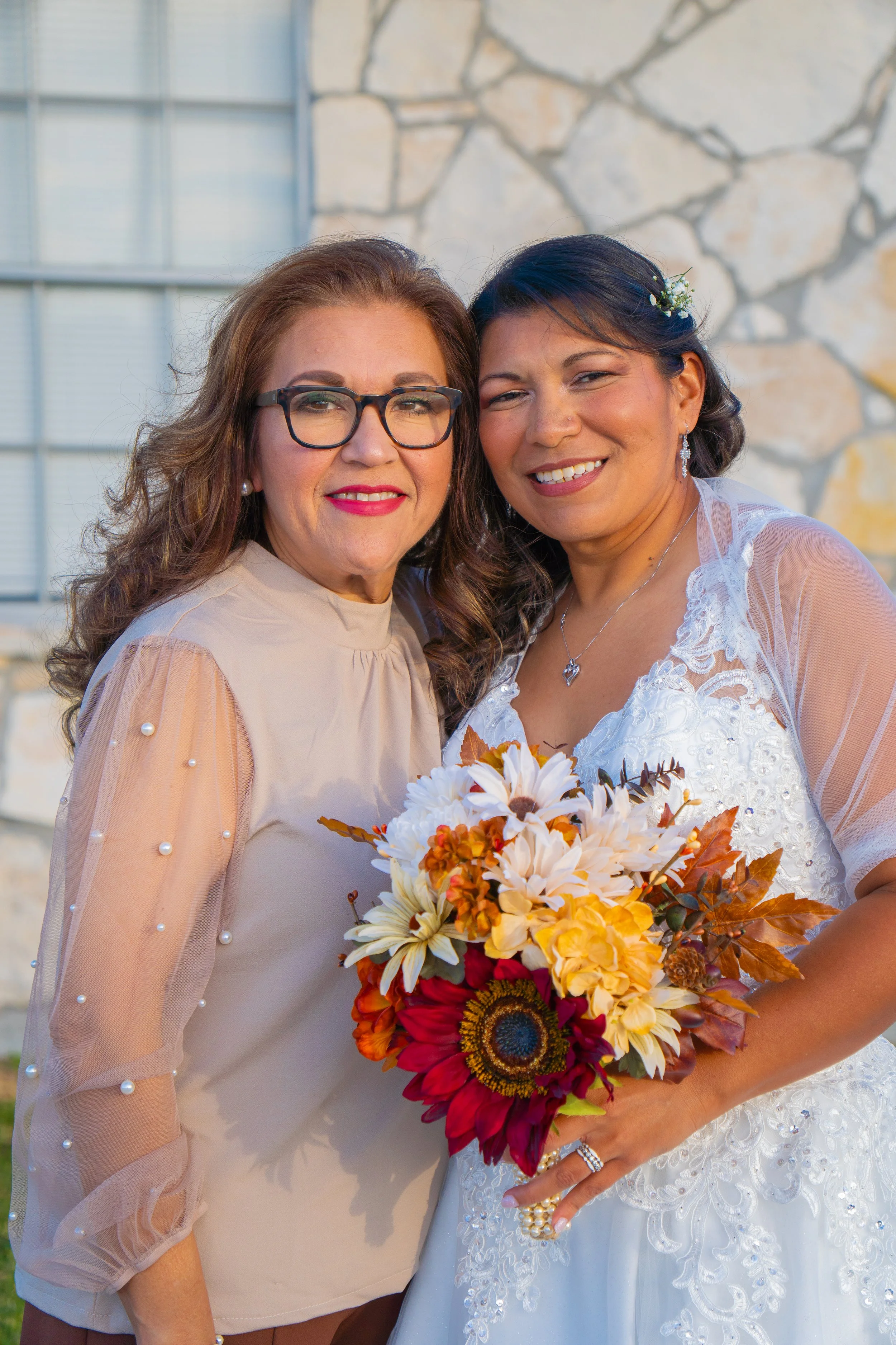 Two women, one in a beige top with pearl accents and glasses, and the other in a white wedding dress holding a bouquet of fall flowers, standing close together and smiling.