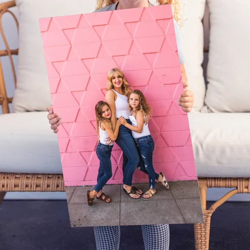 Person holding a framed photo of three girls smiling and hugging against a pink geometric wall.