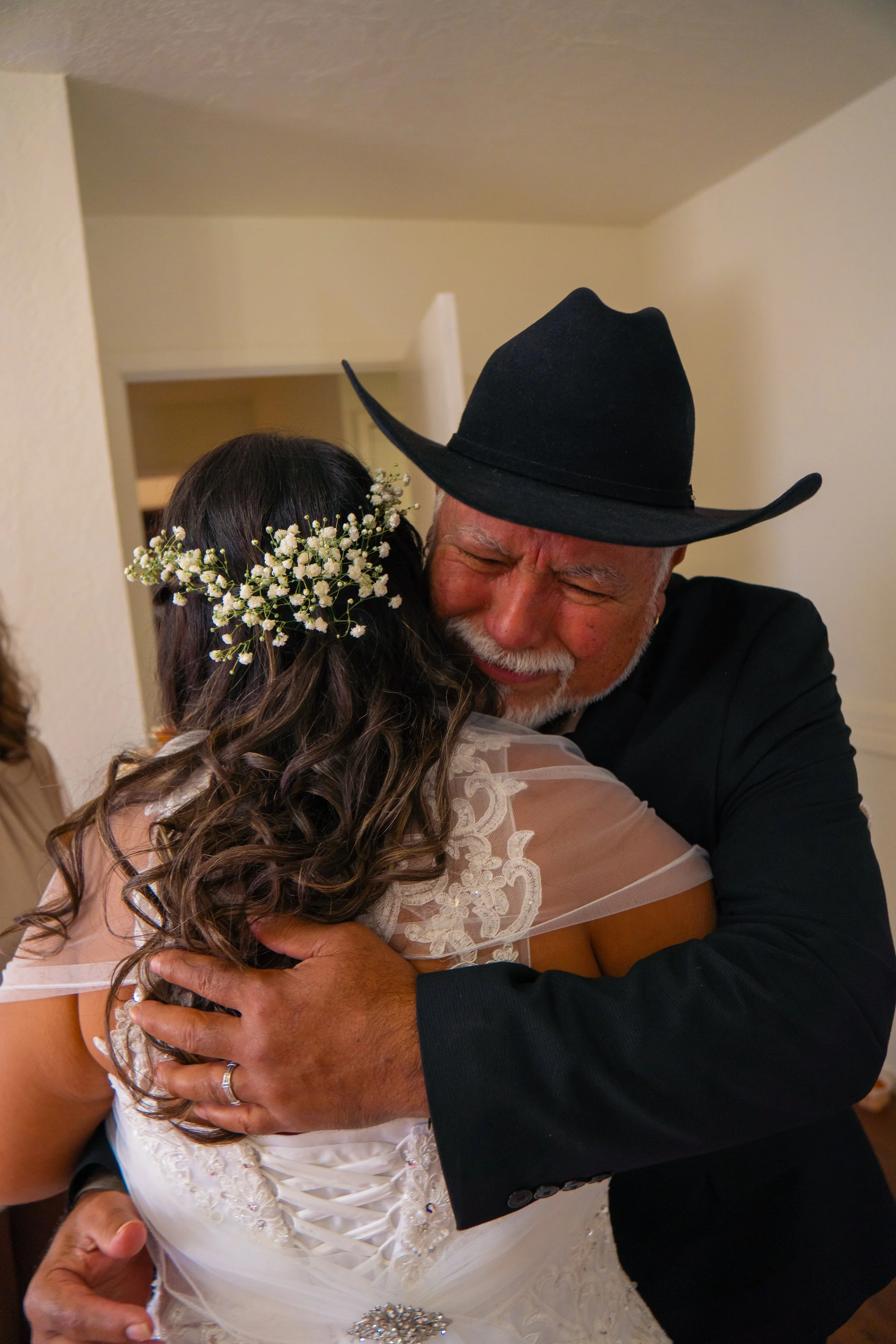 An elderly man wearing a cowboy hat is embracing a woman in a wedding dress. The woman has long, curly hair with small white flowers woven into it, and the man is smiling with his eyes closed.