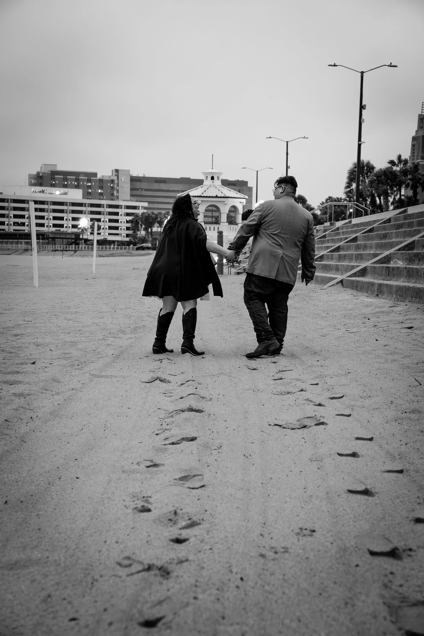 A black and white photo of a couple holding hands while walking on a sandy beach near stairs, city buildings, and palm trees in the background.