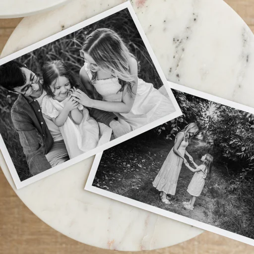 Two printed black-and-white photographs placed on a round surface, one showing a family of four sitting together, and the other of a woman and a girl holding hands outdoors.
