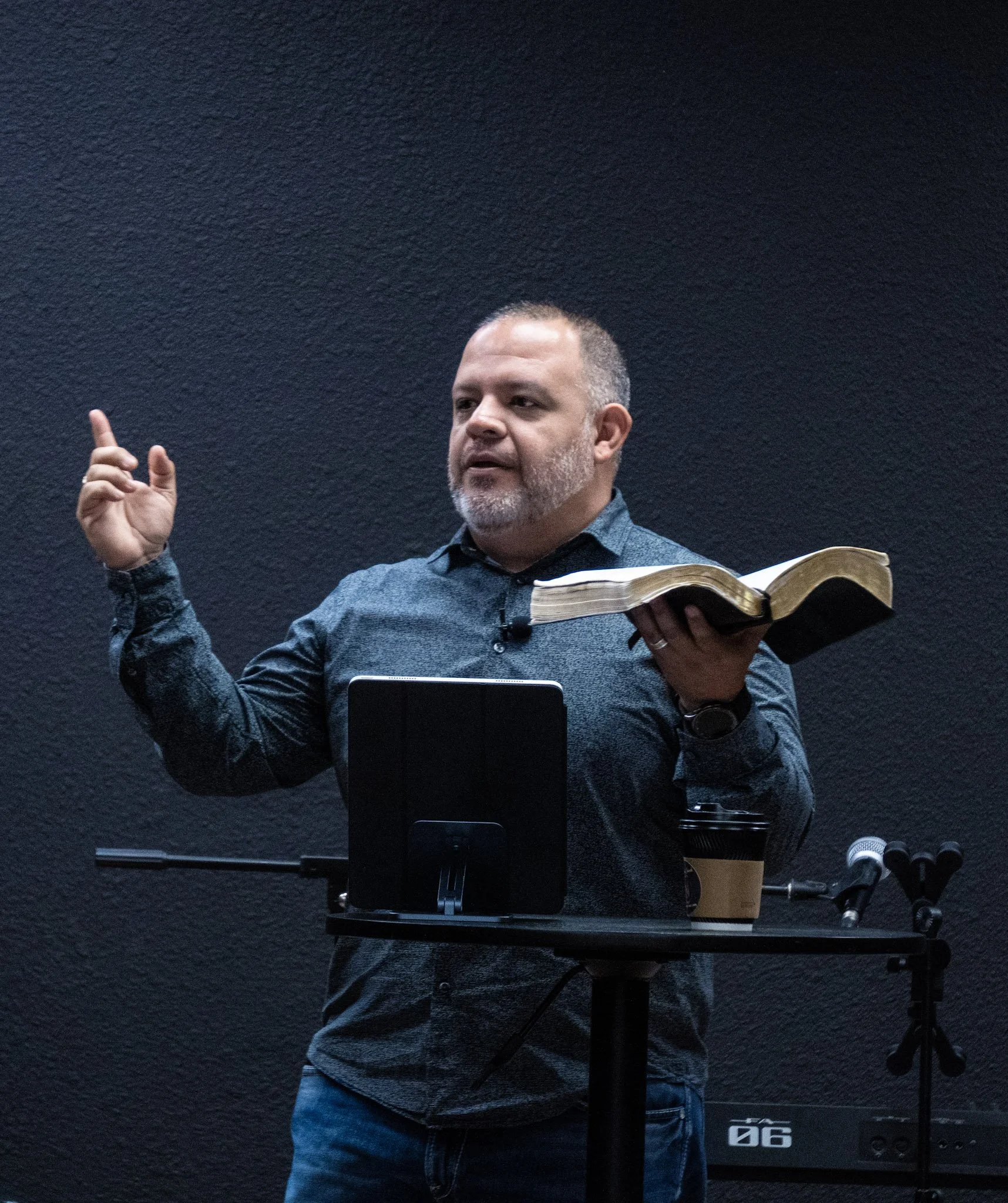 A man with a beard, wearing a dark shirt, speaking with one hand raised and holding an open book in the other, standing at a podium with a tablet, coffee cup, and microphones against a dark textured wall.