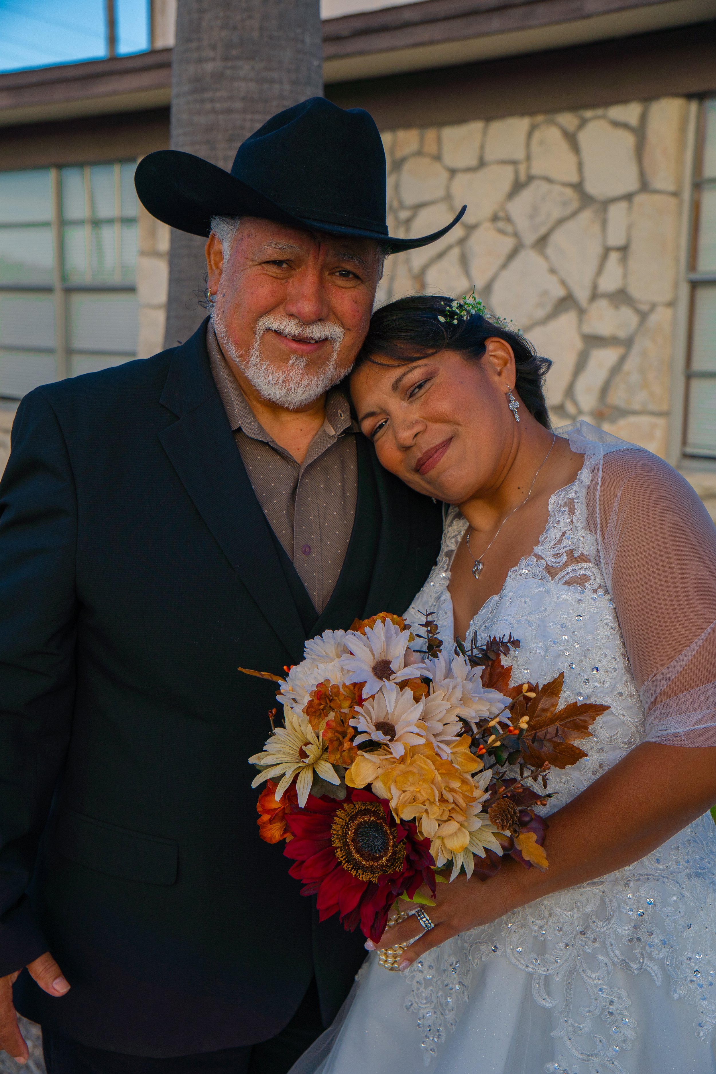 Senior couple on their wedding day, the man in a black suit and cowboy hat, the woman in a white wedding dress holding a bouquet of flowers, standing outdoors near a building with stone and glass windows, smiling and posing closely.