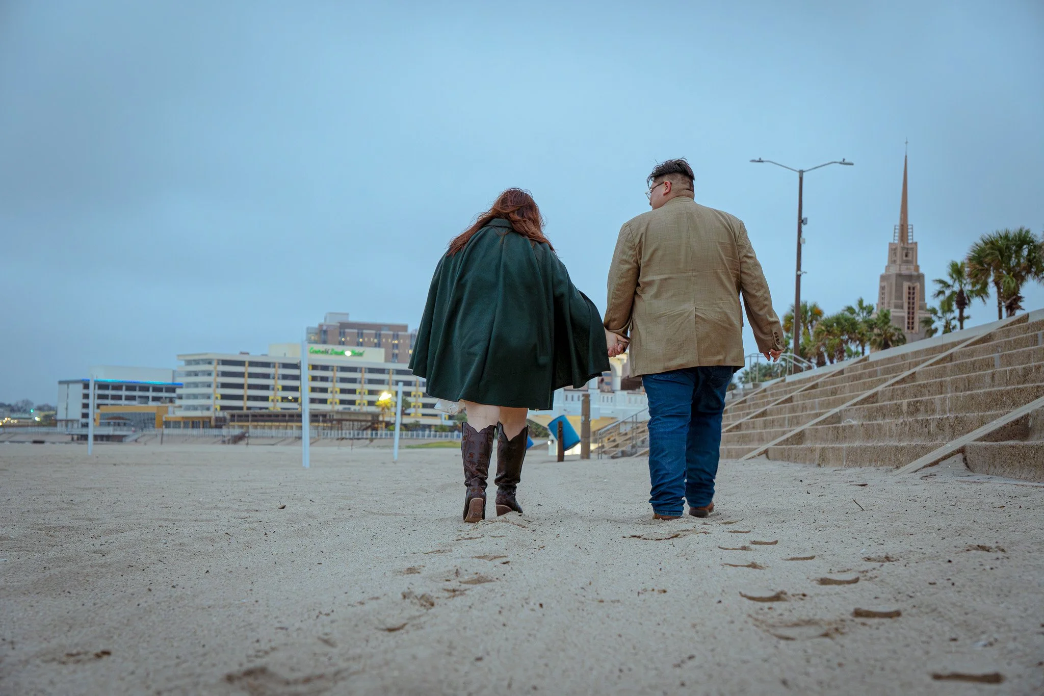 A couple walking hand in hand on a sandy beach, facing away from the camera, during the evening with city buildings and a church steeple in the background.