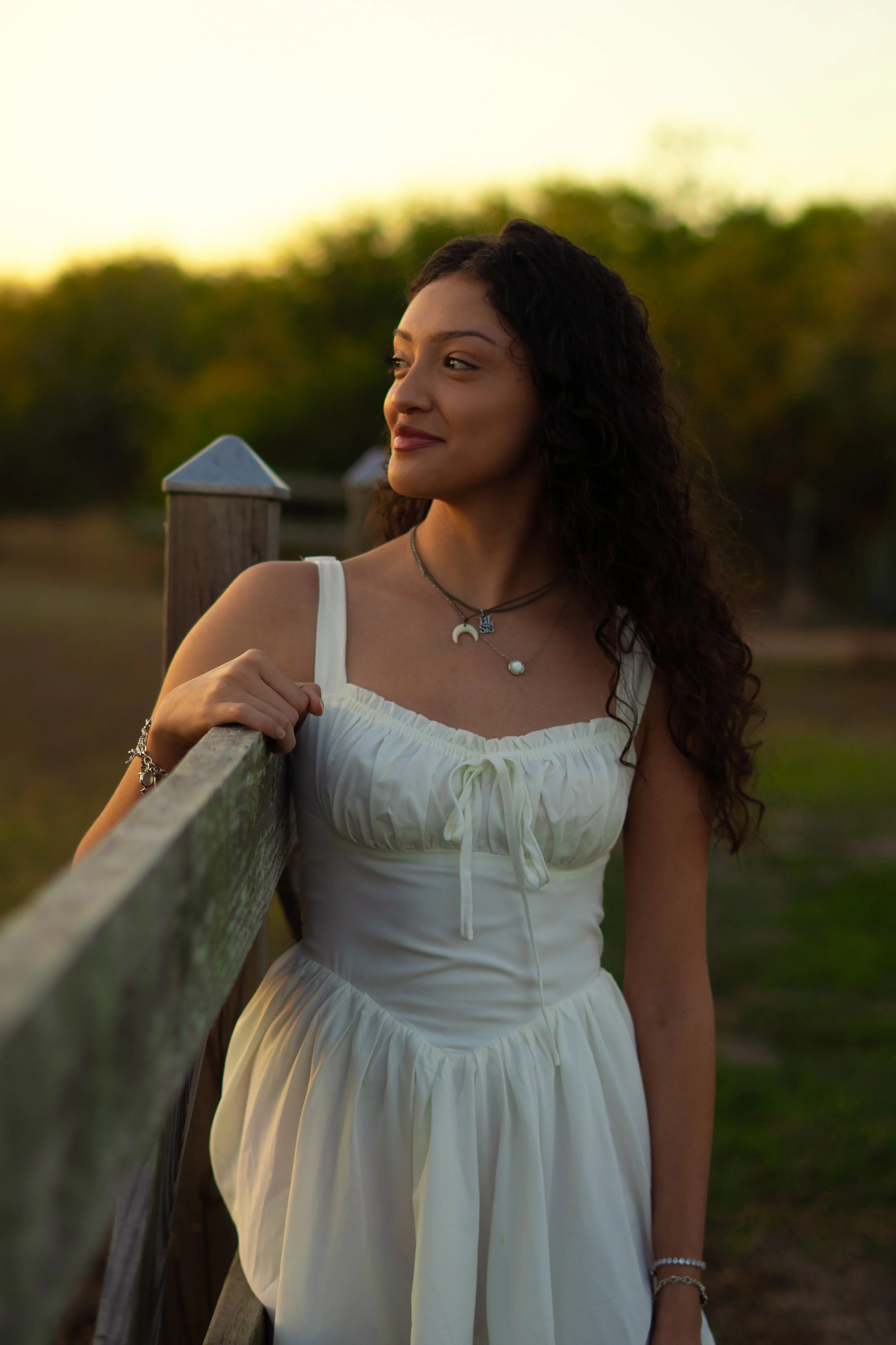 A young woman in a white dress with spaghetti straps, standing outdoors during sunset, leaning on a wooden fence with a blurred green background.