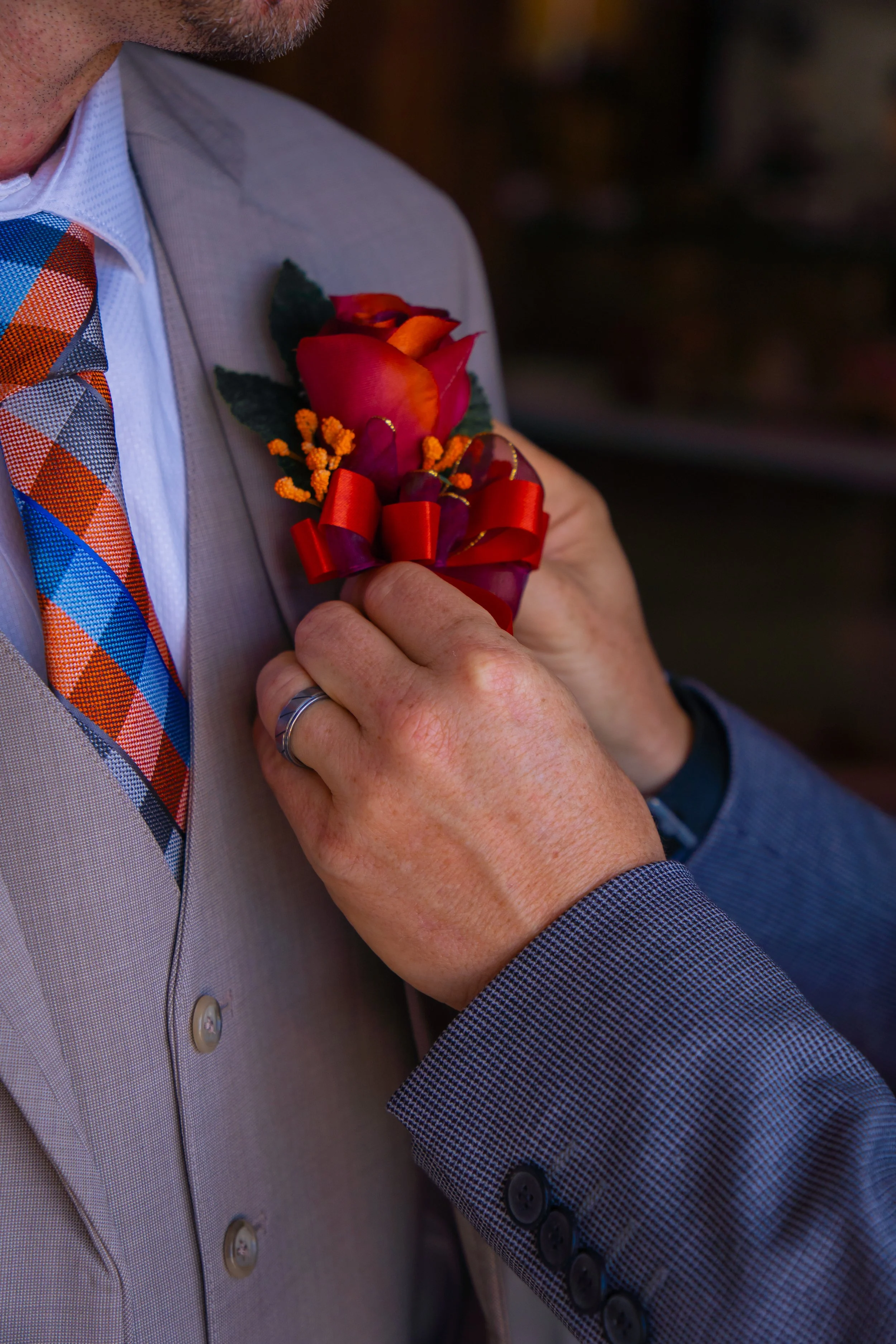 A person is pinning a red and orange boutonniere with yellow accents onto a light gray suit jacket, with a plaid tie and a white dress shirt underneath.