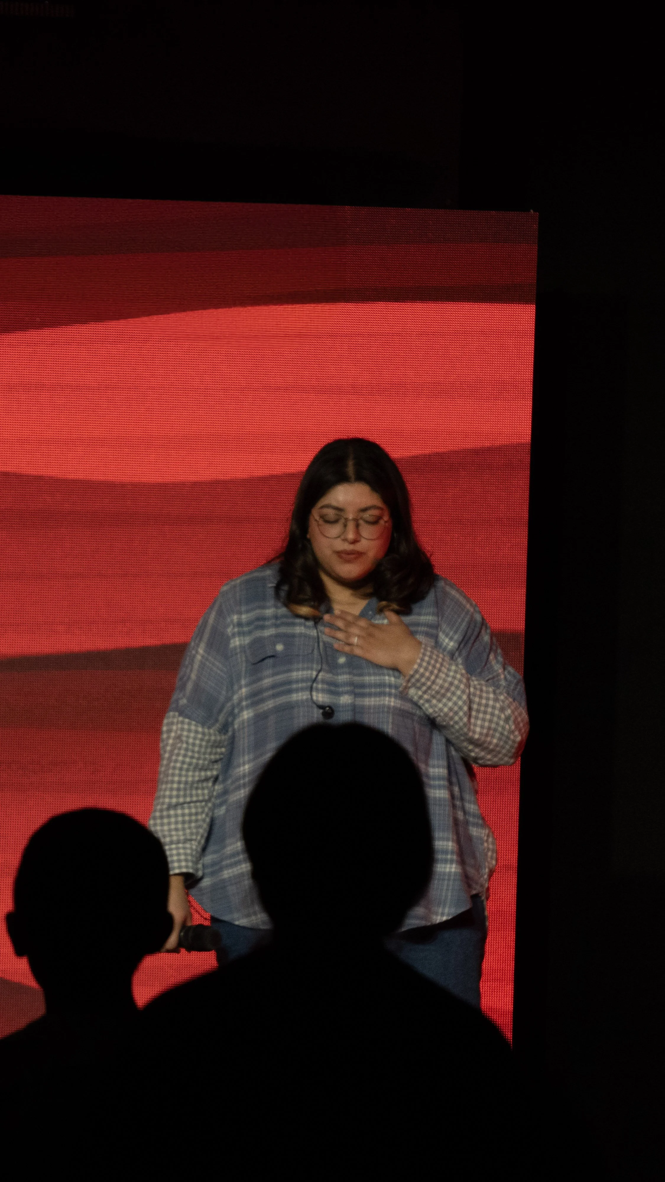 A woman with glasses and dark hair on stage with a red background, holding her hand on her chest, facing the audience.