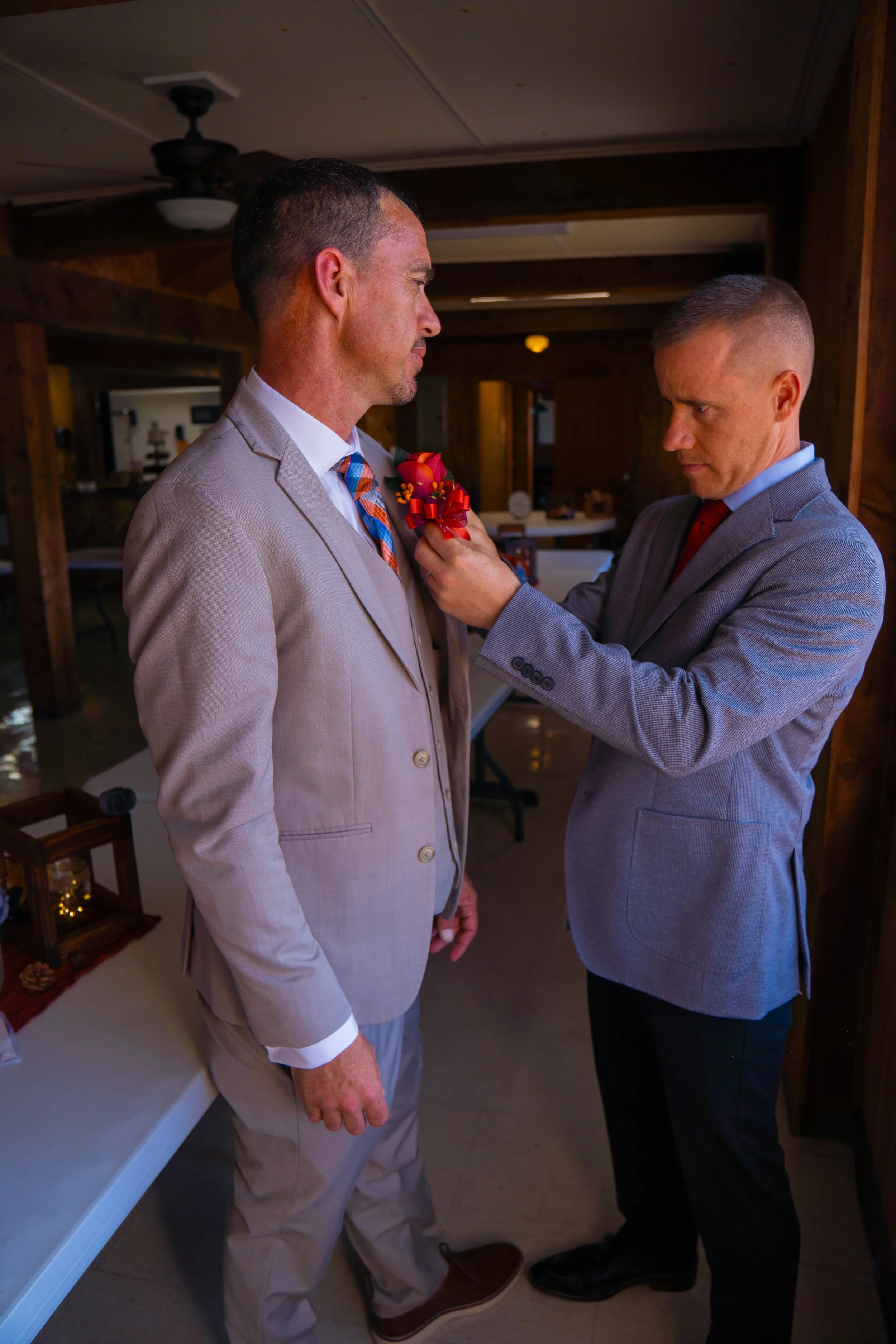 A man in a beige suit is being pinning a red boutonniere onto the lapel of a man in a gray suit. They are standing indoors with dark wooden walls. The man in the gray suit is looking at the boutonniere with a focused expression. The setting suggests 