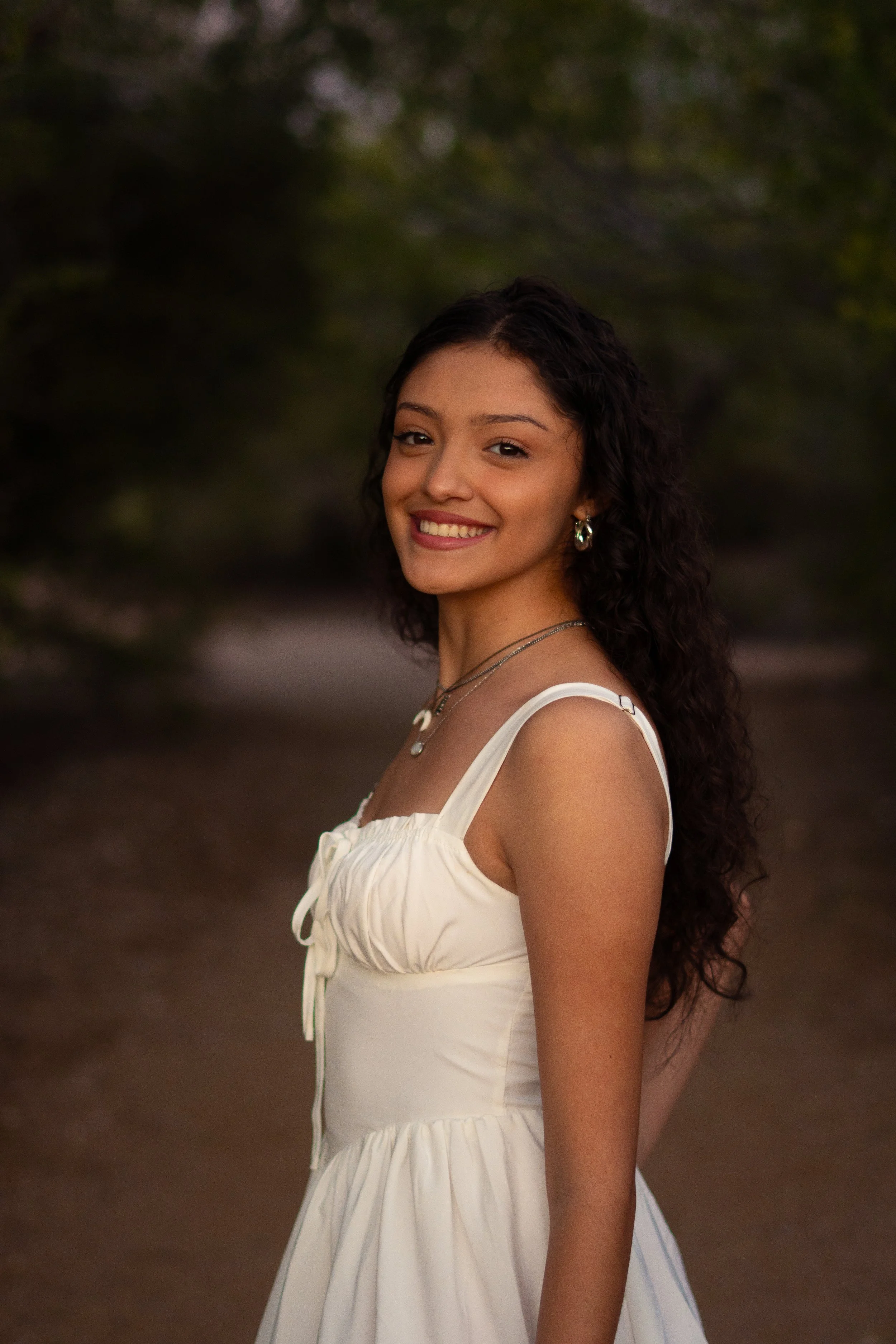 Young woman with dark, curly hair smiling outdoors at sunset, wearing a white dress and jewelry.