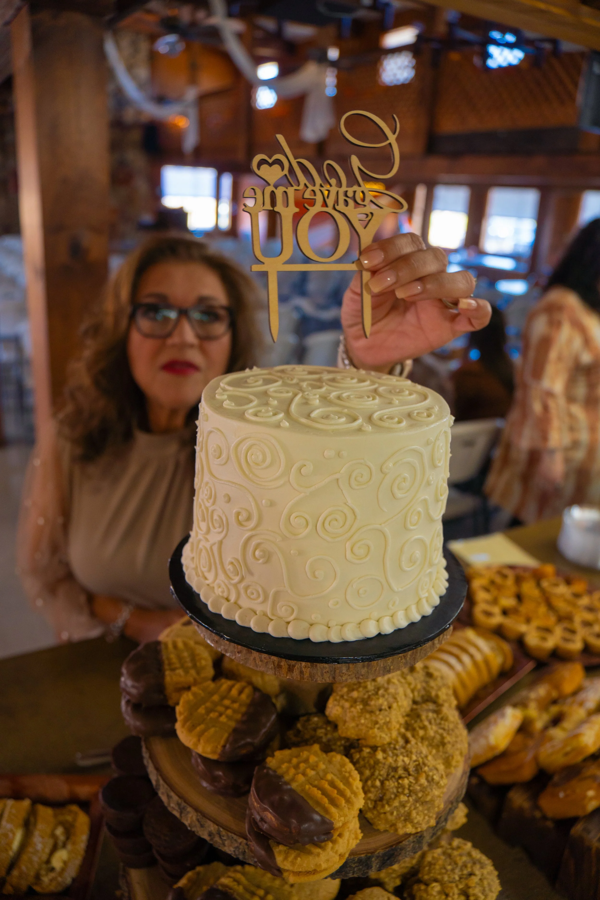 A tiered cake with a swirl pattern, being presented at a celebration. The person holding the cake is slightly blurry in the background, with glasses and wavy hair. The top of the cake has a decoration that says 'happy anniversary to you' in cursive s