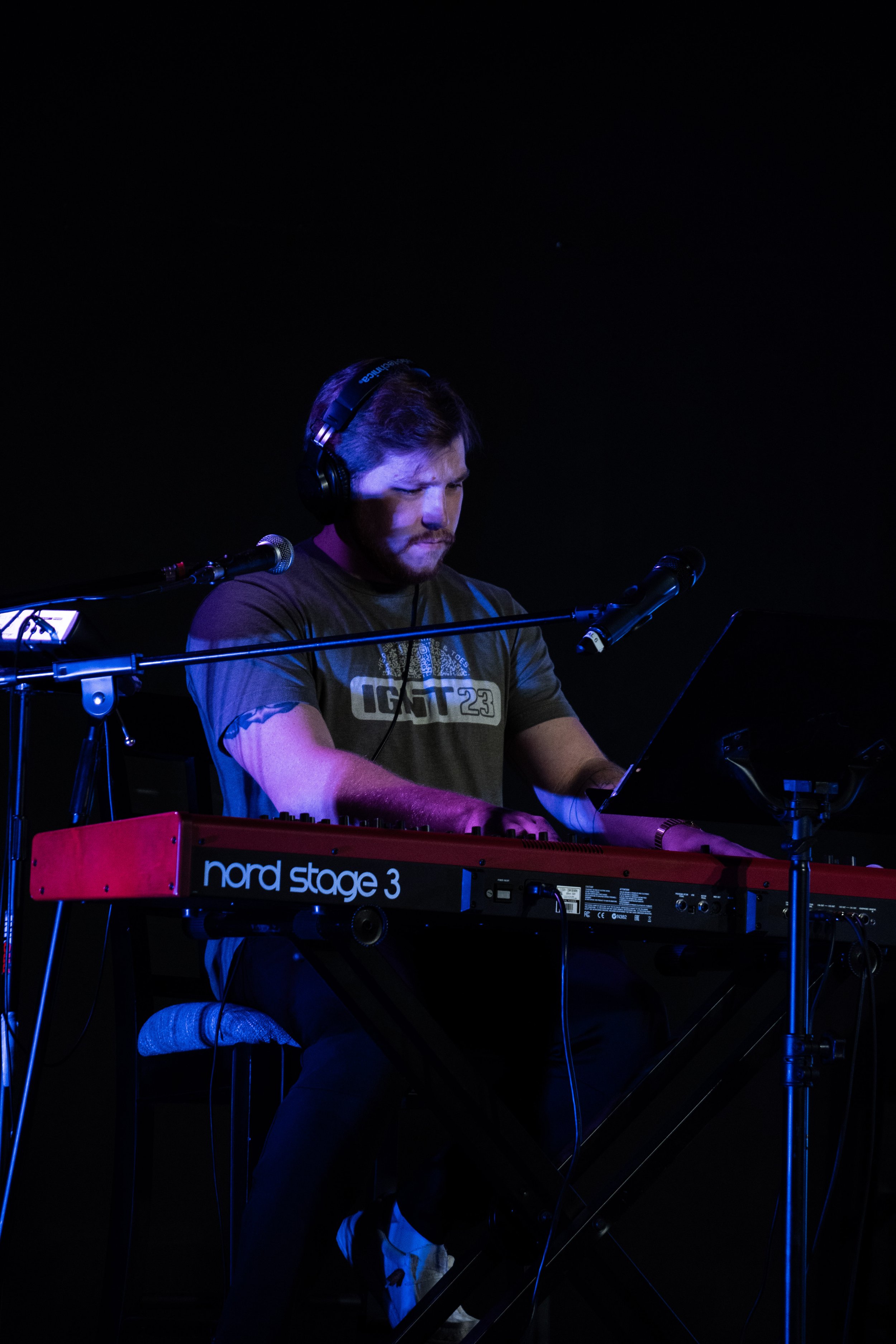 A man wearing headphones is playing a keyboard on stage, with a dark background and two microphones nearby.