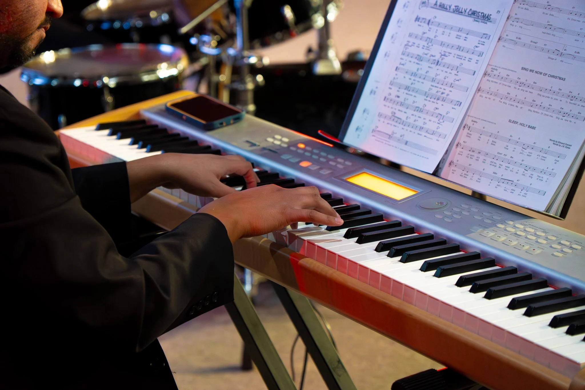 A person playing a keyboard with sheet music on a stand, surrounded by musical instruments and equipment.