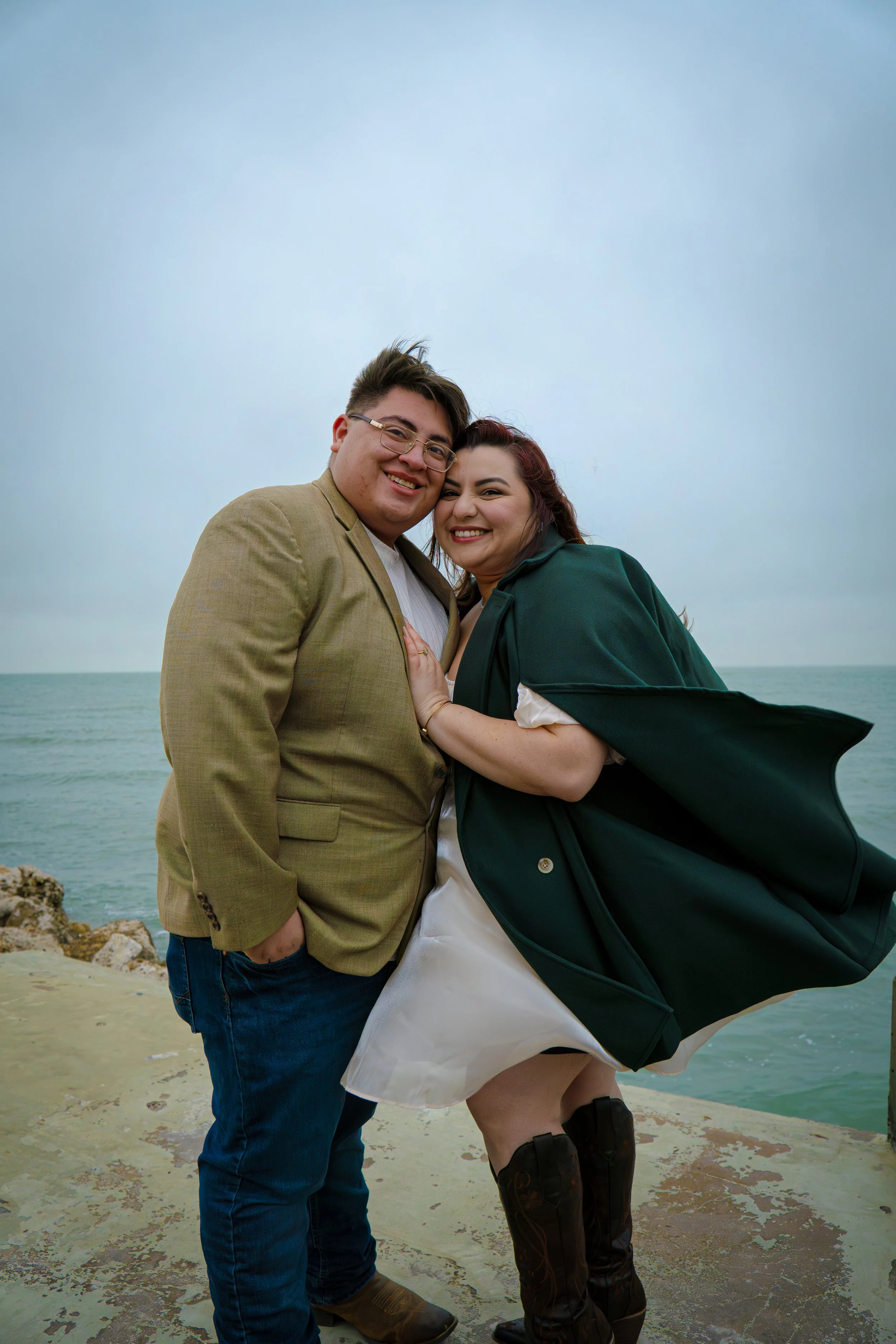 A smiling couple standing close together on a beach, with the sea and cloudy sky in the background. The man is wearing glasses, a beige blazer, and jeans, and the woman is wearing a dark green cape, a white dress, and black knee-high boots.