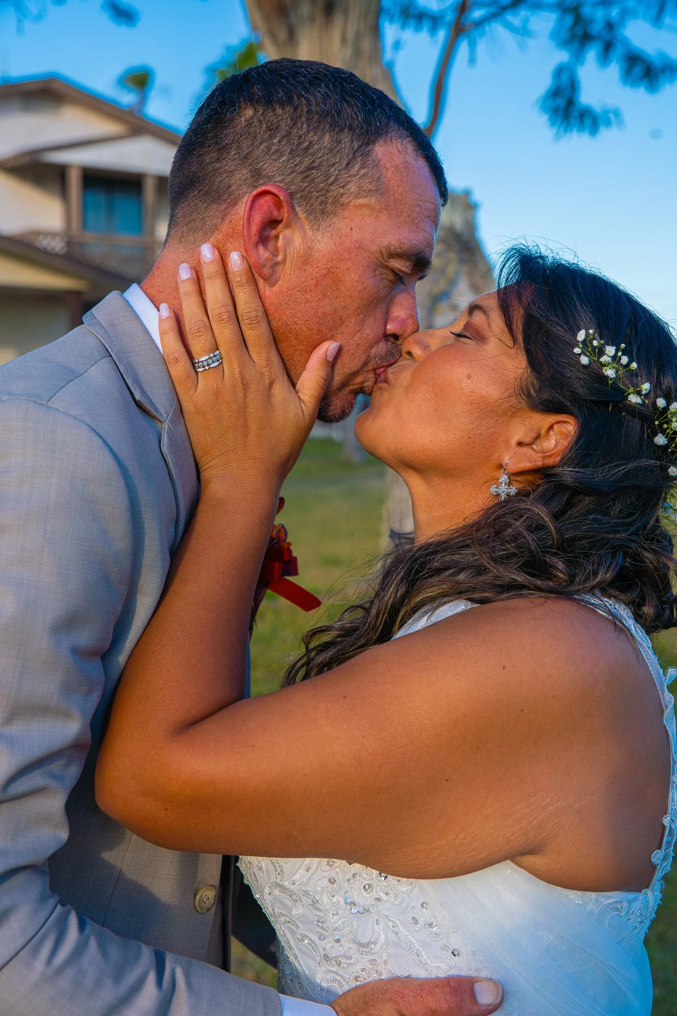 A couple in wedding attire sharing a kiss outdoors during sunset, with trees and a house in the background.