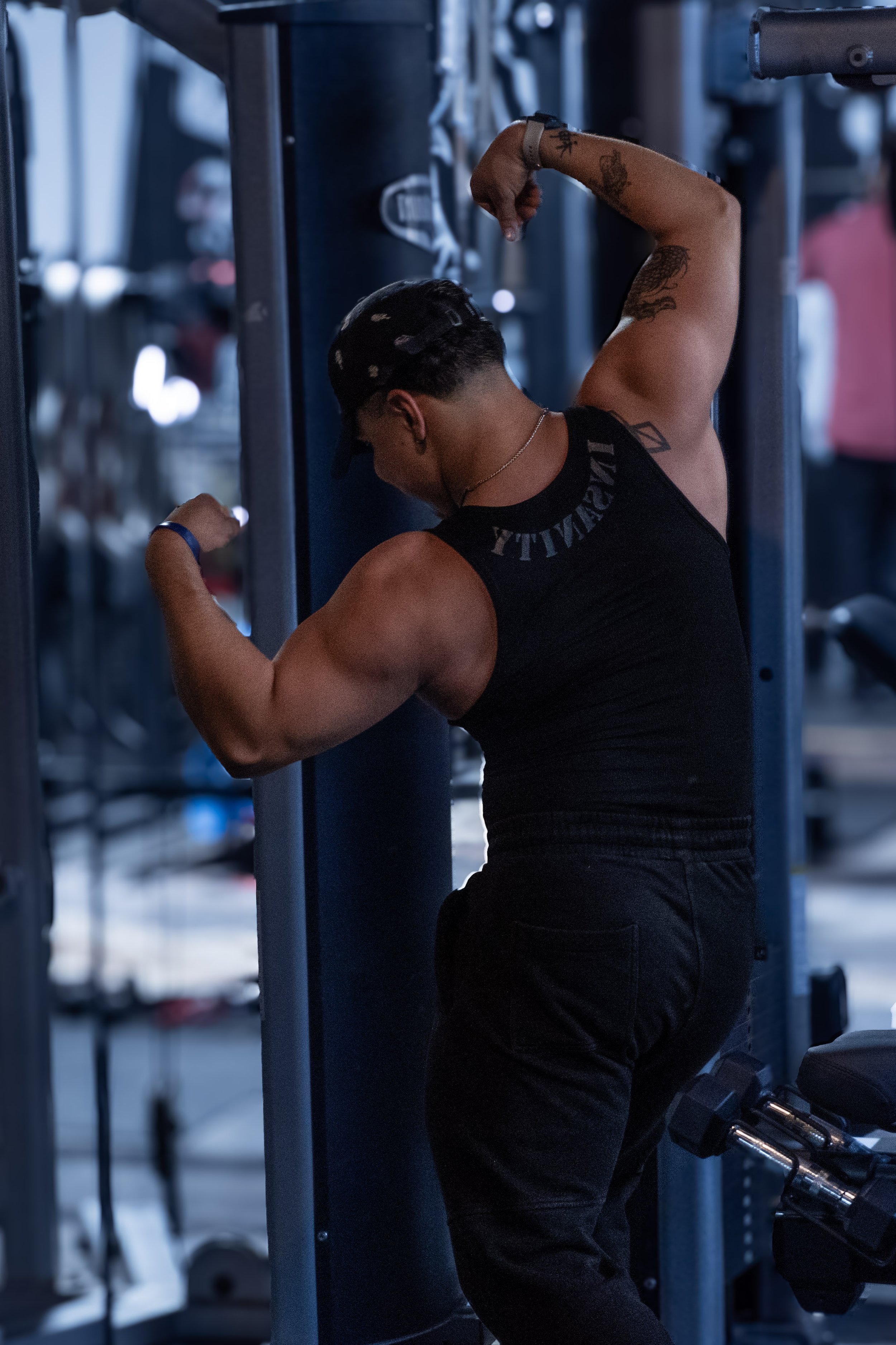 A muscular man with tattoos on his arms, wearing a black tank top and black pants, is performing a bicep curl at the gym, flexing his right arm against a workout equipment.
