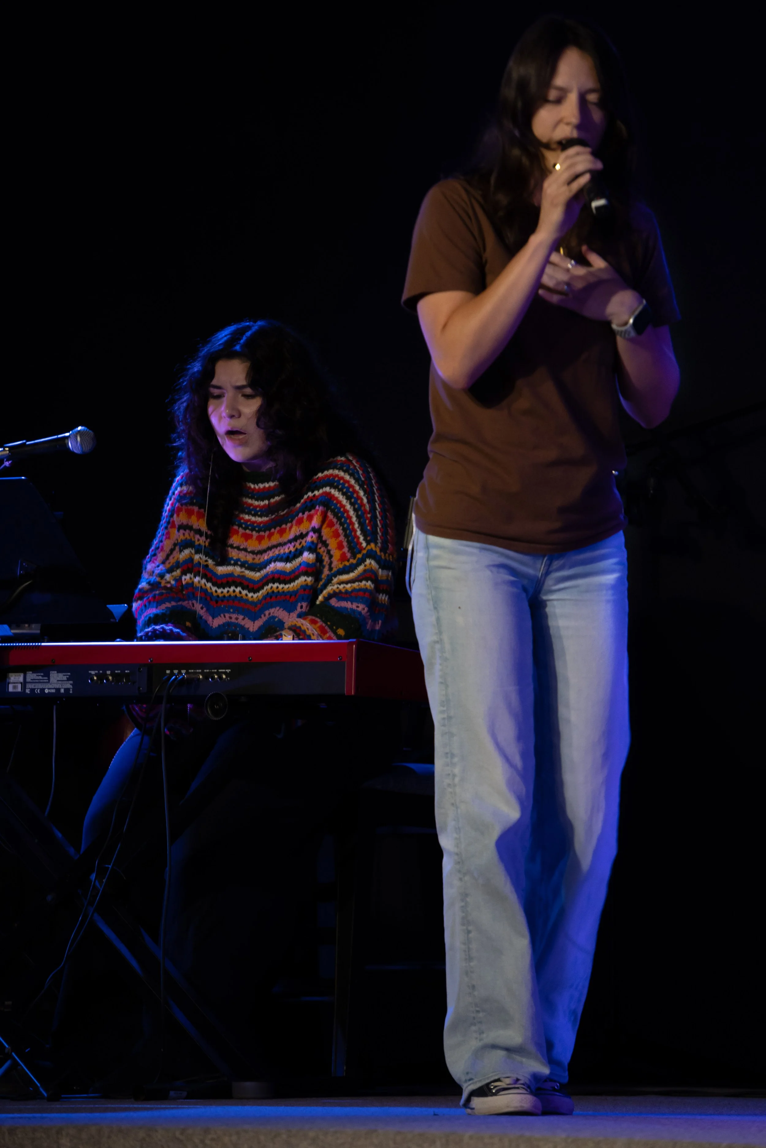 Two women performing on stage; one is singing into a microphone with eyes closed and the other is playing the keyboard, wearing a colorful striped sweater.