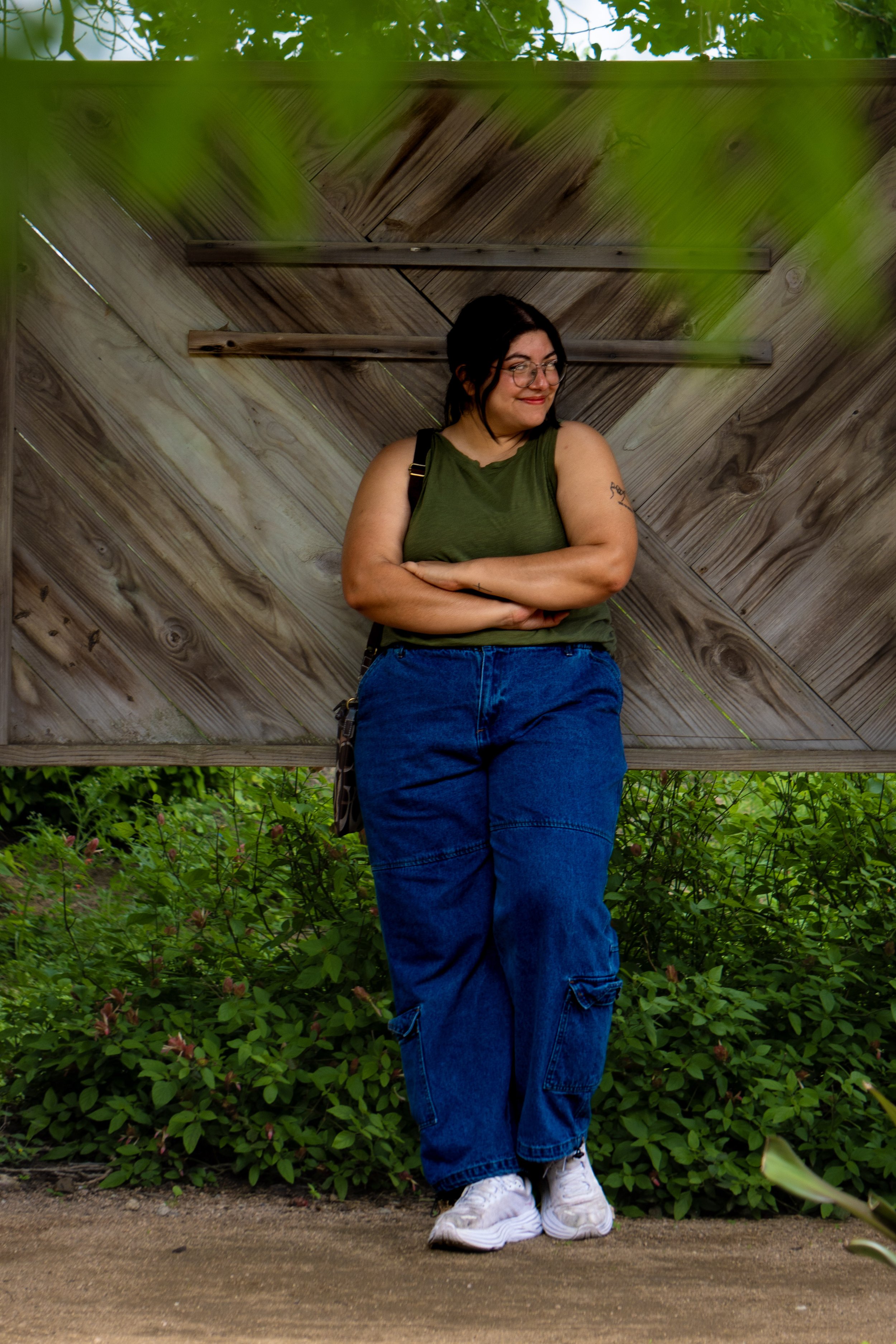 A woman with glasses and dark hair styled in a bun, wearing a green sleeveless top, baggy blue jeans, and white sneakers, stands with arms crossed in front of a wooden fence and lush green bushes, smiling softly.