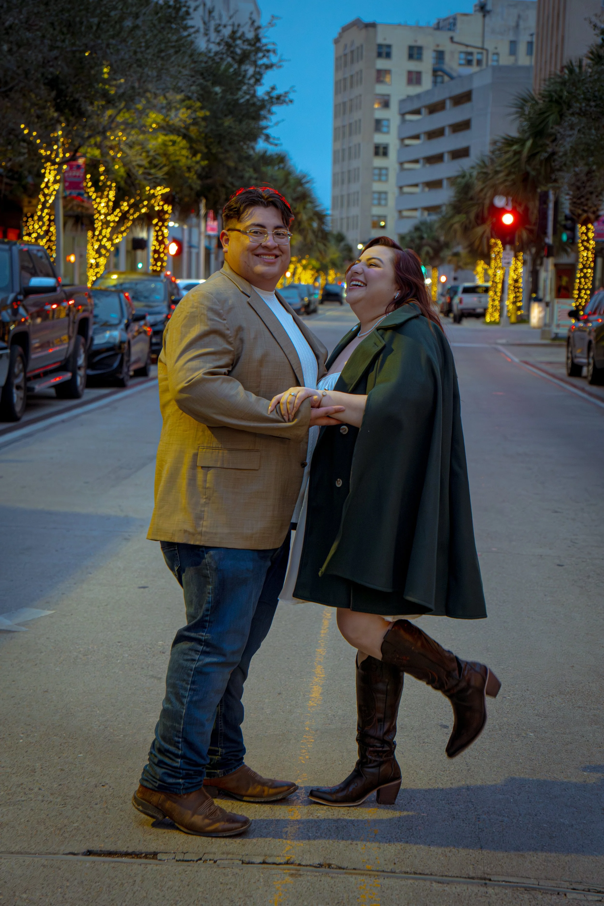 A smiling man and woman holding hands on a city street at dusk, with festive string lights on trees and buildings in the background. The woman is wearing a dark green jacket, a white dress, and knee-high brown boots with heels. The man is wearing a t