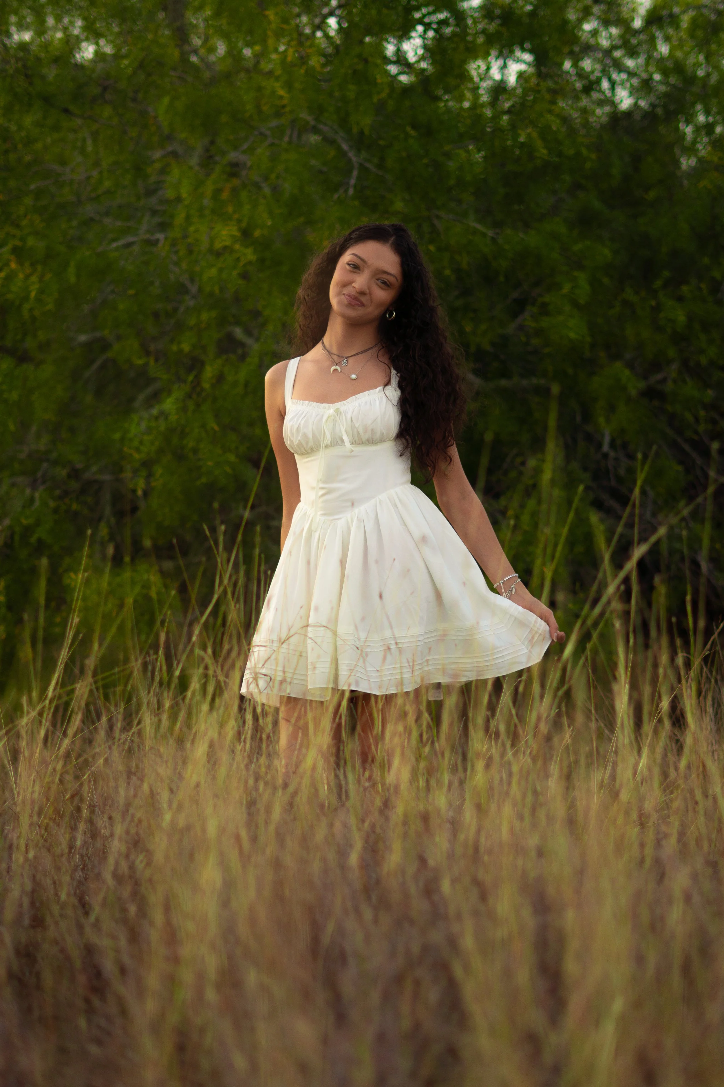 Young woman with long curly hair wearing a white dress and jewelry standing in a field of tall grass with trees in background.