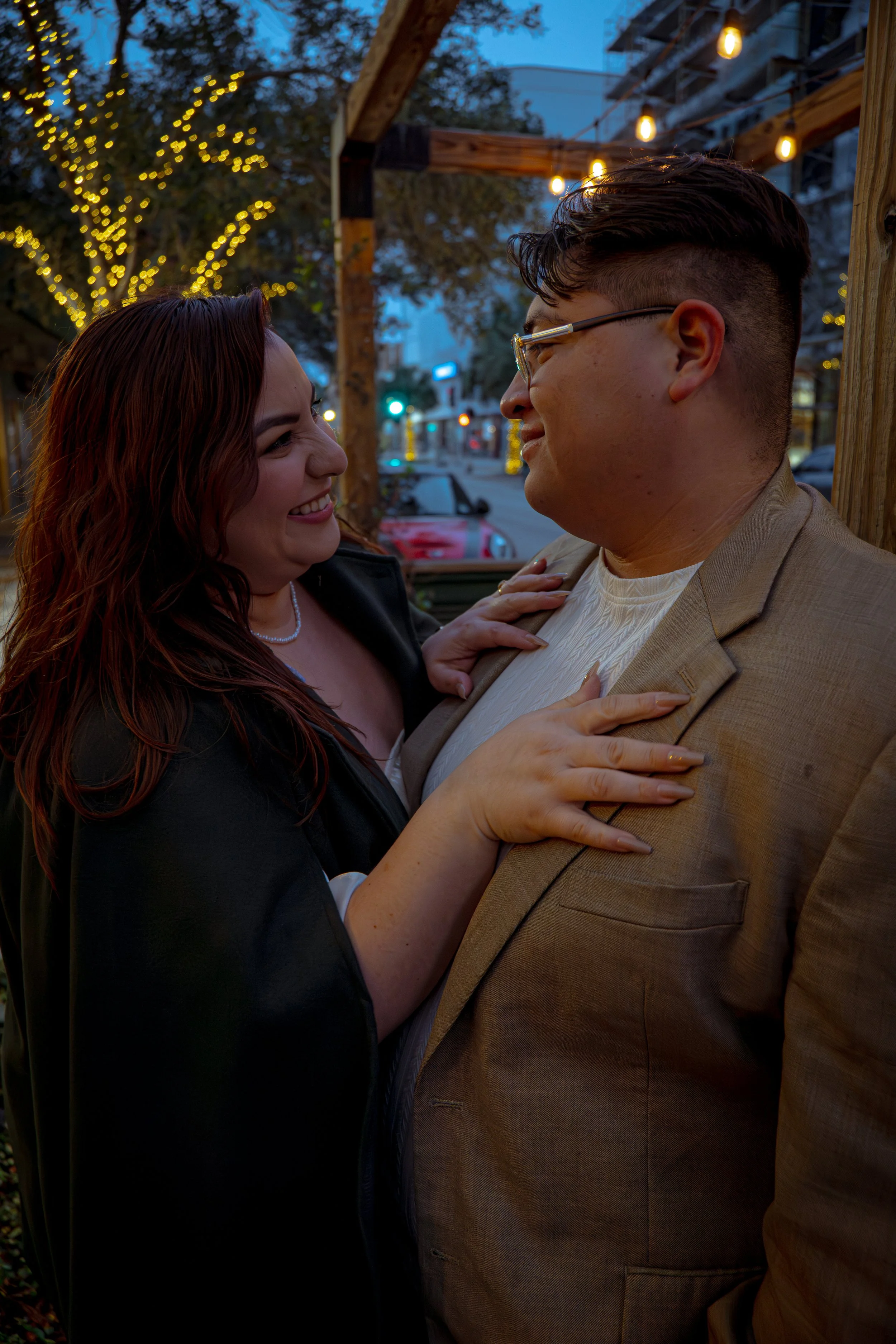 A couple is smiling and looking at each other affectionately, standing close together outdoors during evening time, with string lights and trees illuminated in the background.