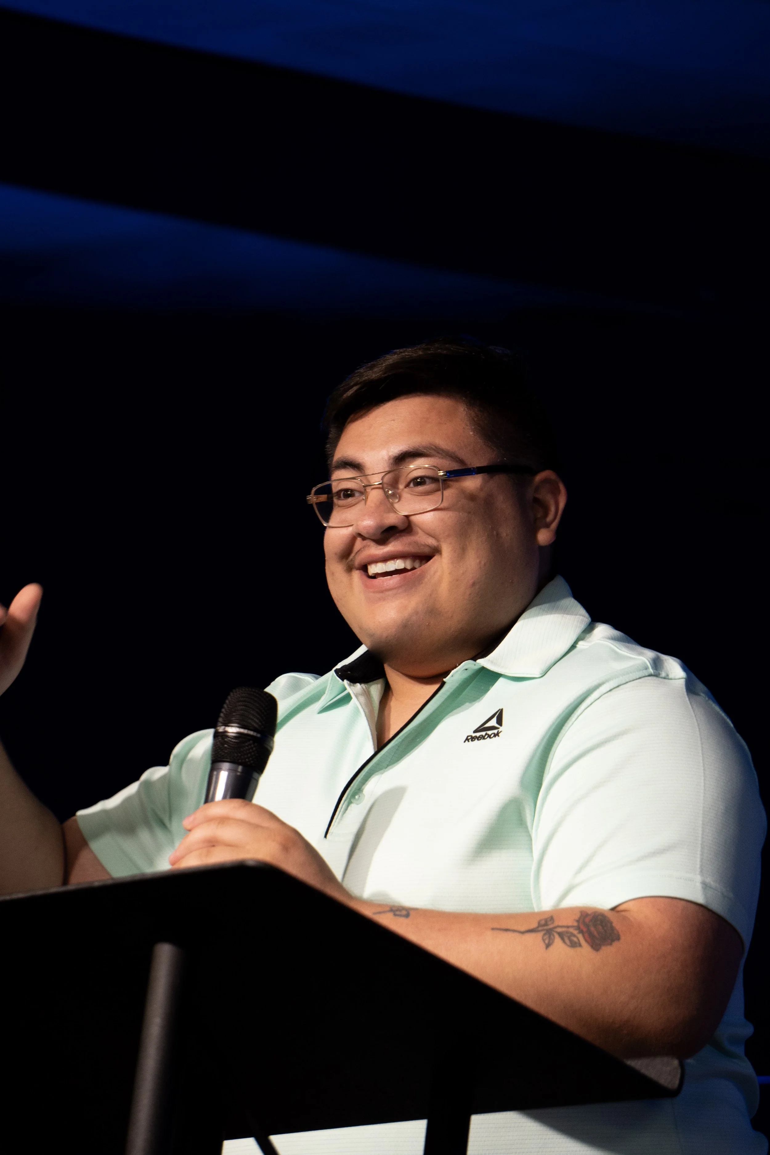 A man wearing glasses and a Reebok polo shirt is standing behind a lectern, smiling, and holding a microphone, gesturing with his other hand, on a stage with a dark background and blue lighting.
