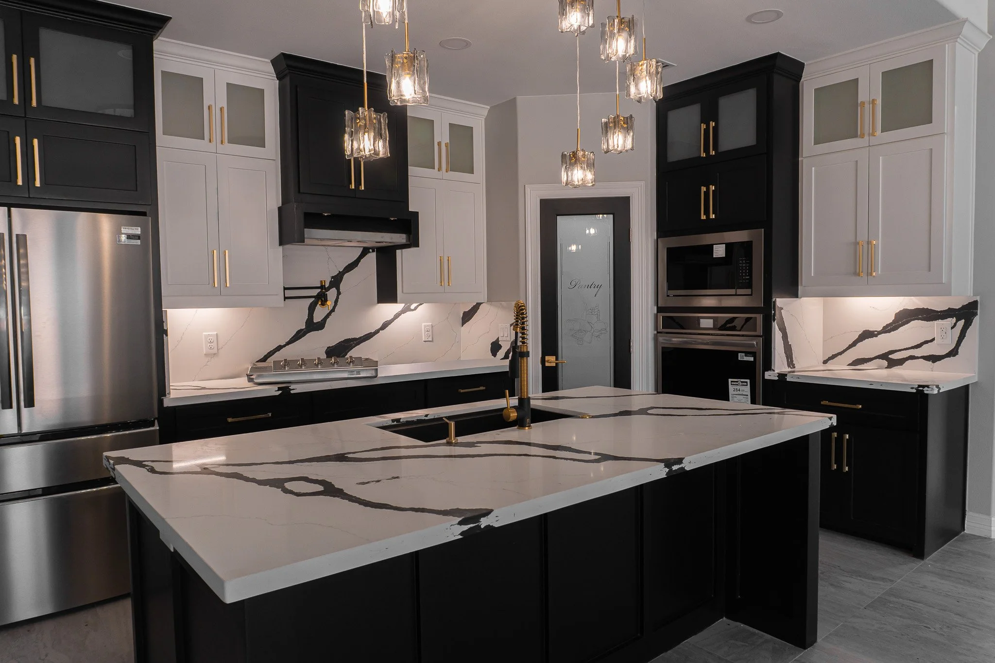 Modern kitchen with black and white cabinetry, marble countertops, and gold accents on cabinet handles and faucet. Features stainless steel appliances, pendant lighting, and a kitchen island.