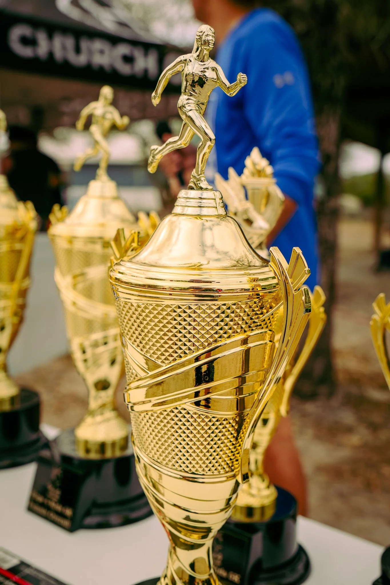 Golden trophy with a figure of a running person on top, displayed outdoors with other trophies and a person in a blue shirt in the background.