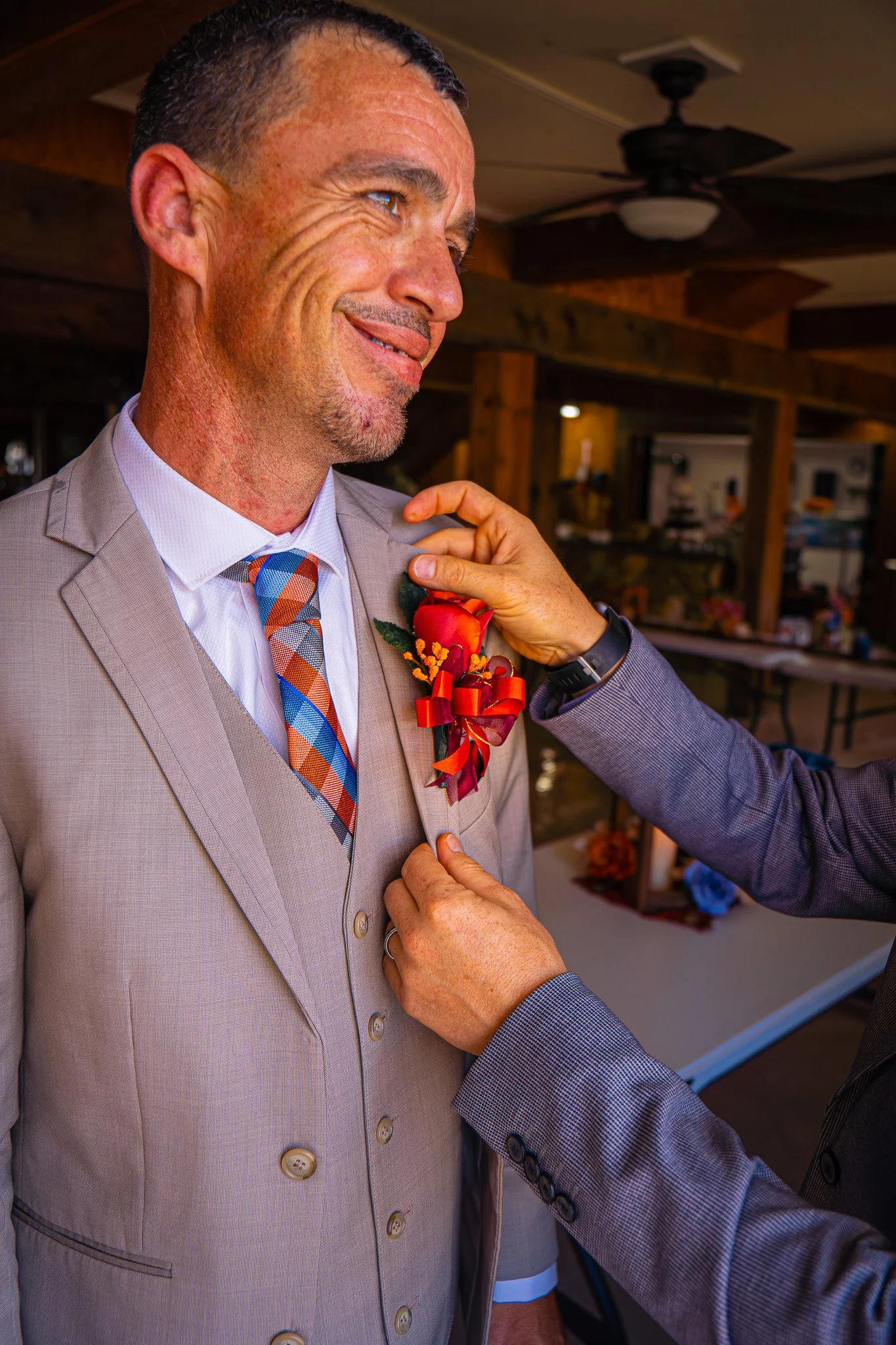 A man in a light gray suit with a colorful plaid tie is having a red boutonniere with orange and yellow flowers pinned to his lapel by another person. The man is smiling and looking slightly to the side. The background appears to be a rustic indoor s