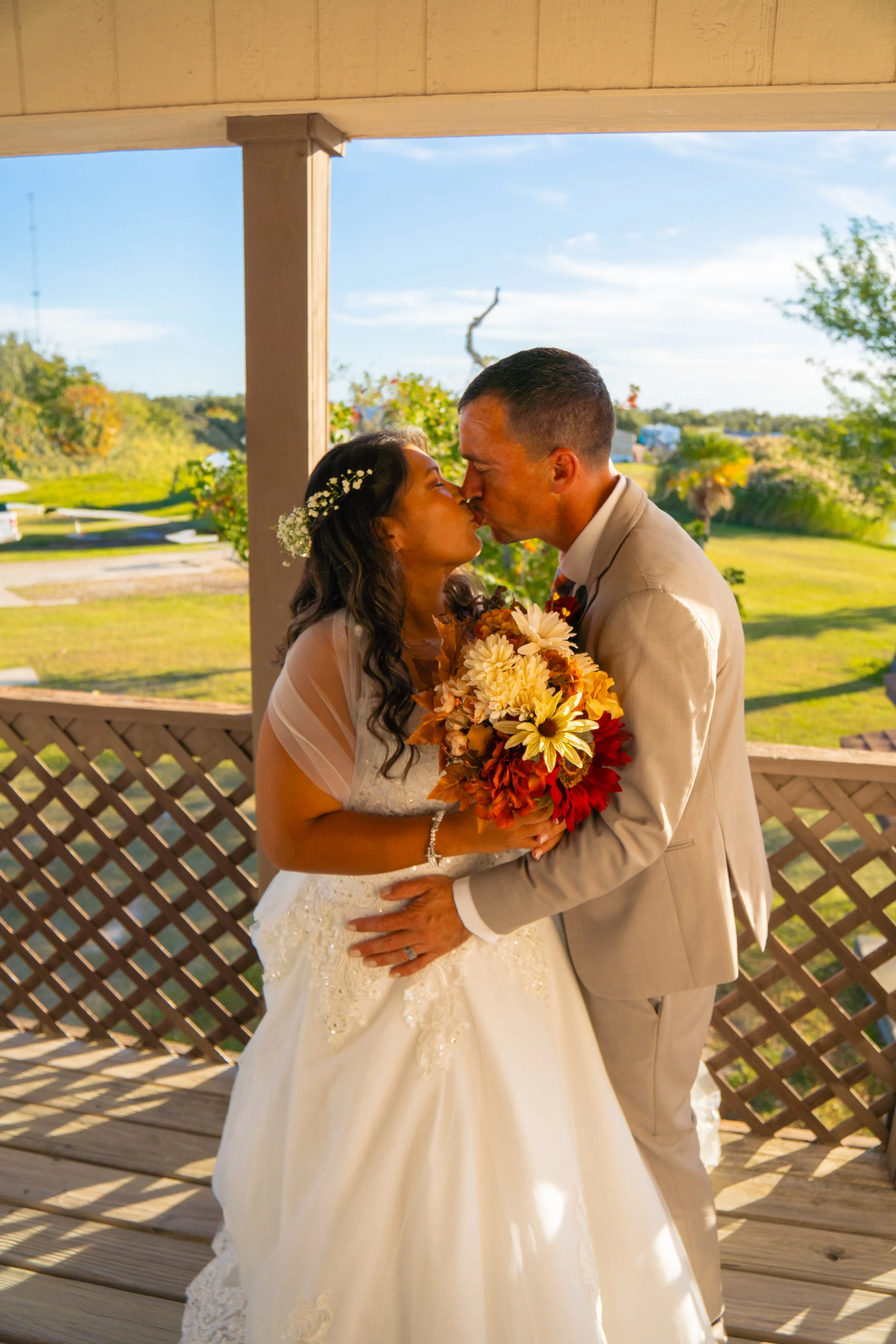 A bride and groom kissing outdoors, with the bride holding a large bouquet of flowers. The bride has dark hair with a floral accessory, and the groom has short dark hair, wearing a tan suit. They are standing under a wooden structure, with greenery a