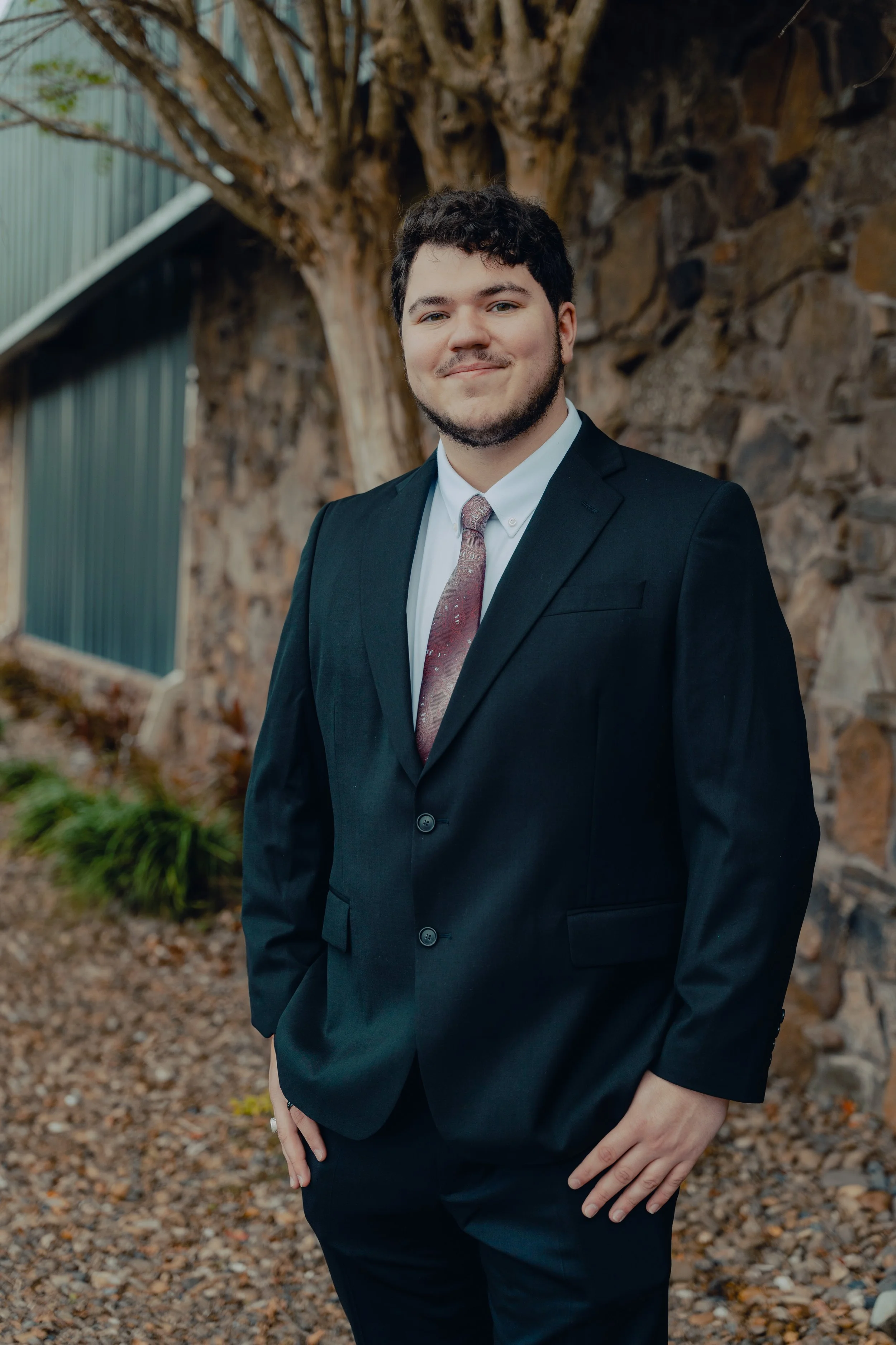 A young man in a black suit, white shirt, and burgundy tie standing outdoors in front of a stone wall and a large tree.