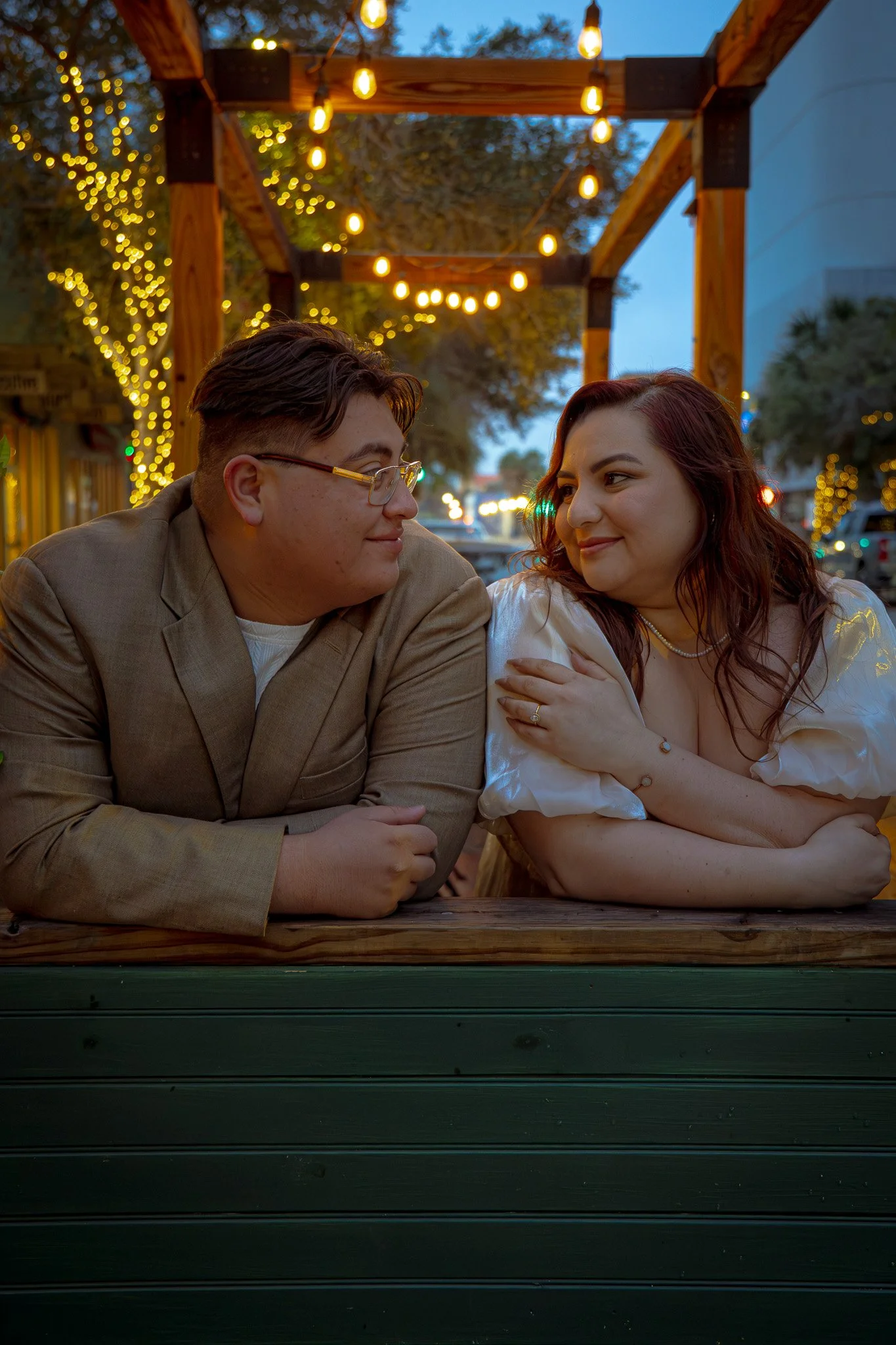 A man and a woman sit at an outdoor table during evening with string lights overhead, gazing into each other's eyes in a cozy, warmly lit setting.