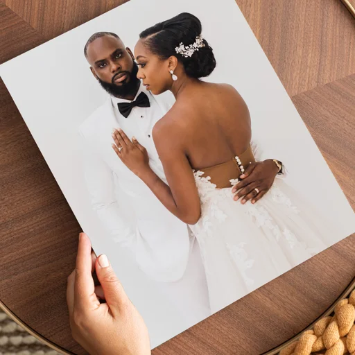A couple in wedding attire posing for a portrait. The groom is in a white tuxedo with a black bow tie, and the bride is in a white strapless wedding gown with lace details, wearing a hair accessory and earrings.