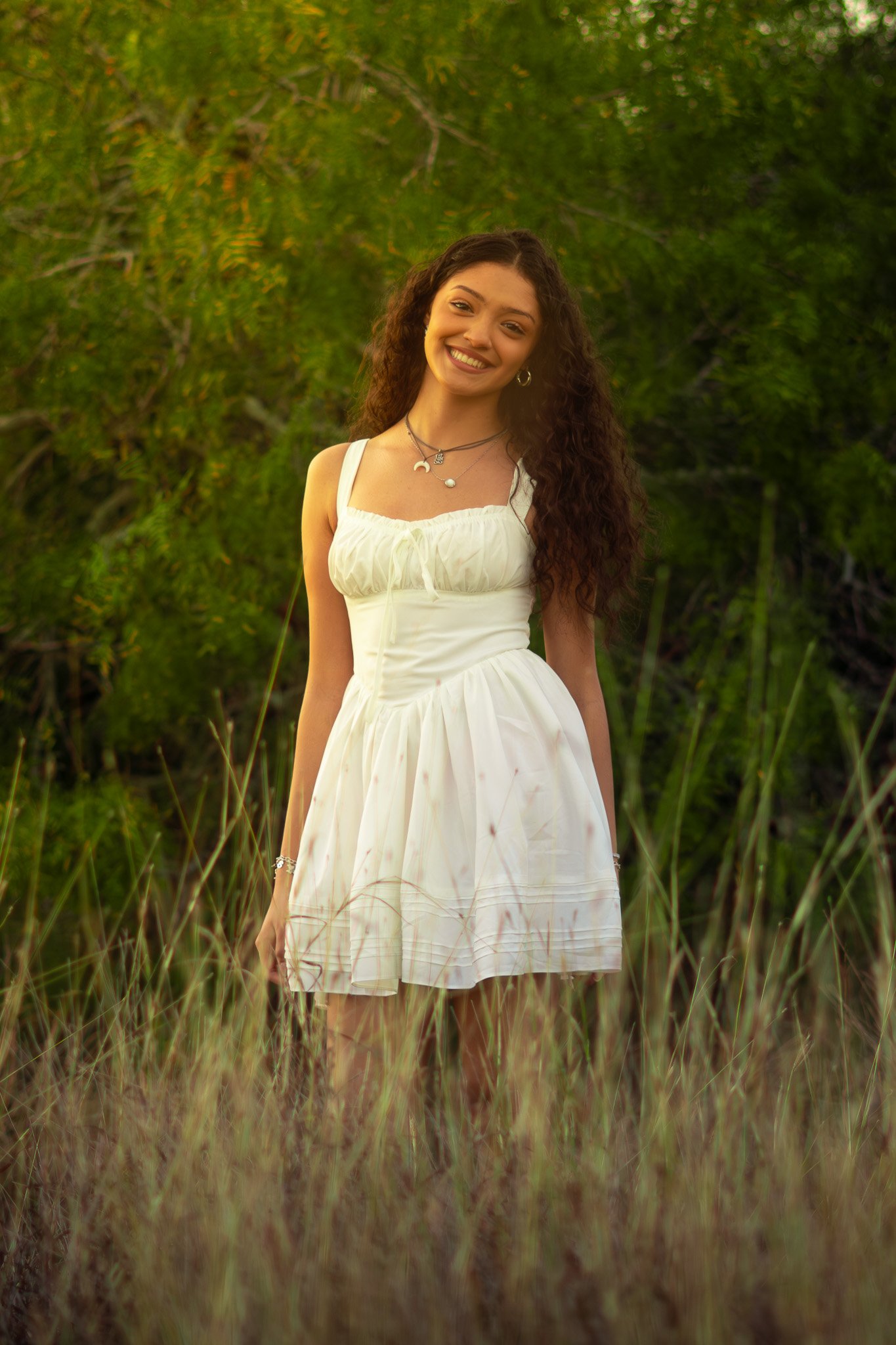 A young woman with long curly hair smiling in a white sleeveless dress standing outdoors surrounded by tall grass and green trees.