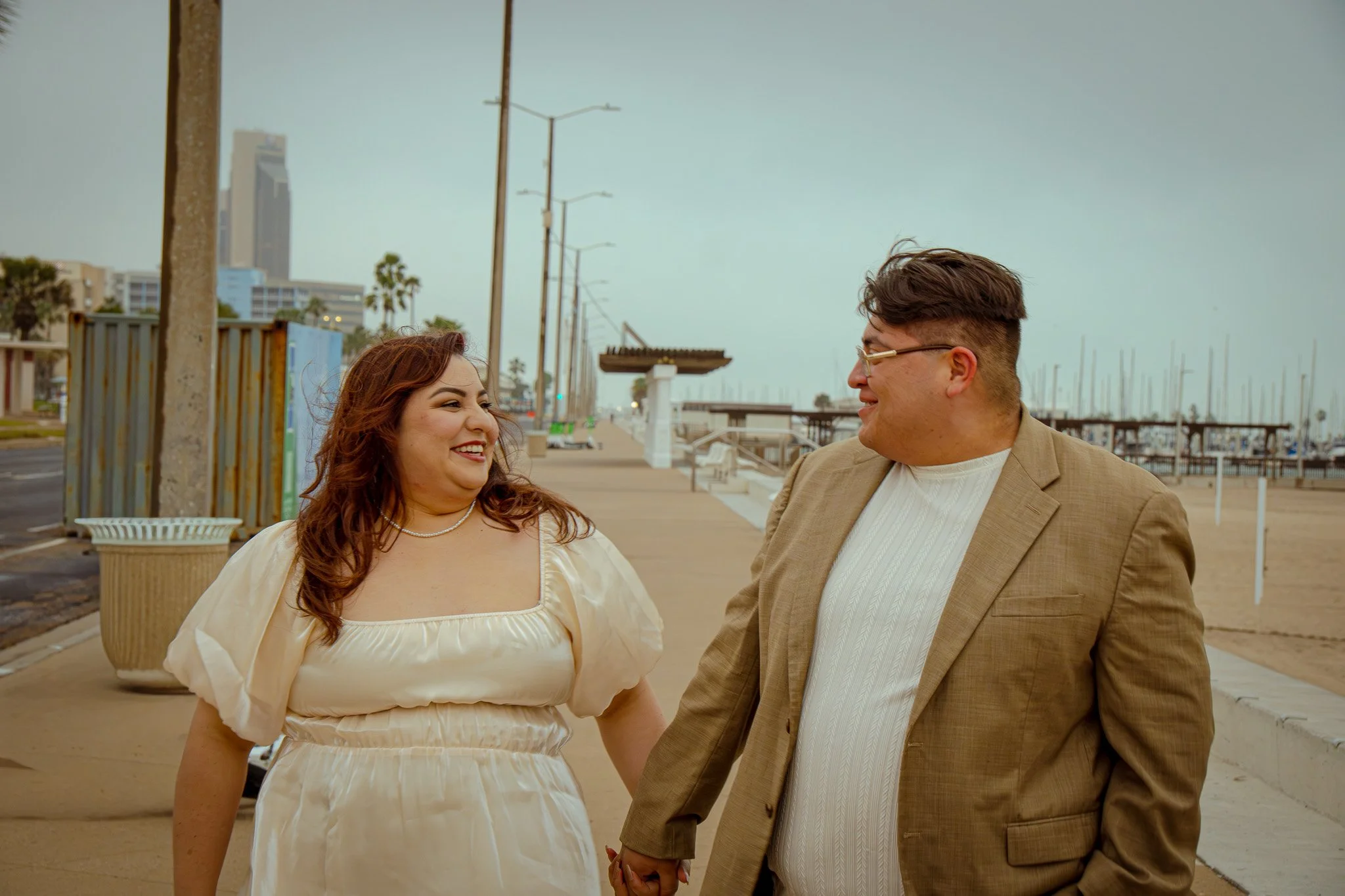 A couple holding hands and smiling at each other on a sidewalk near a beach, with palm trees and buildings in the background.