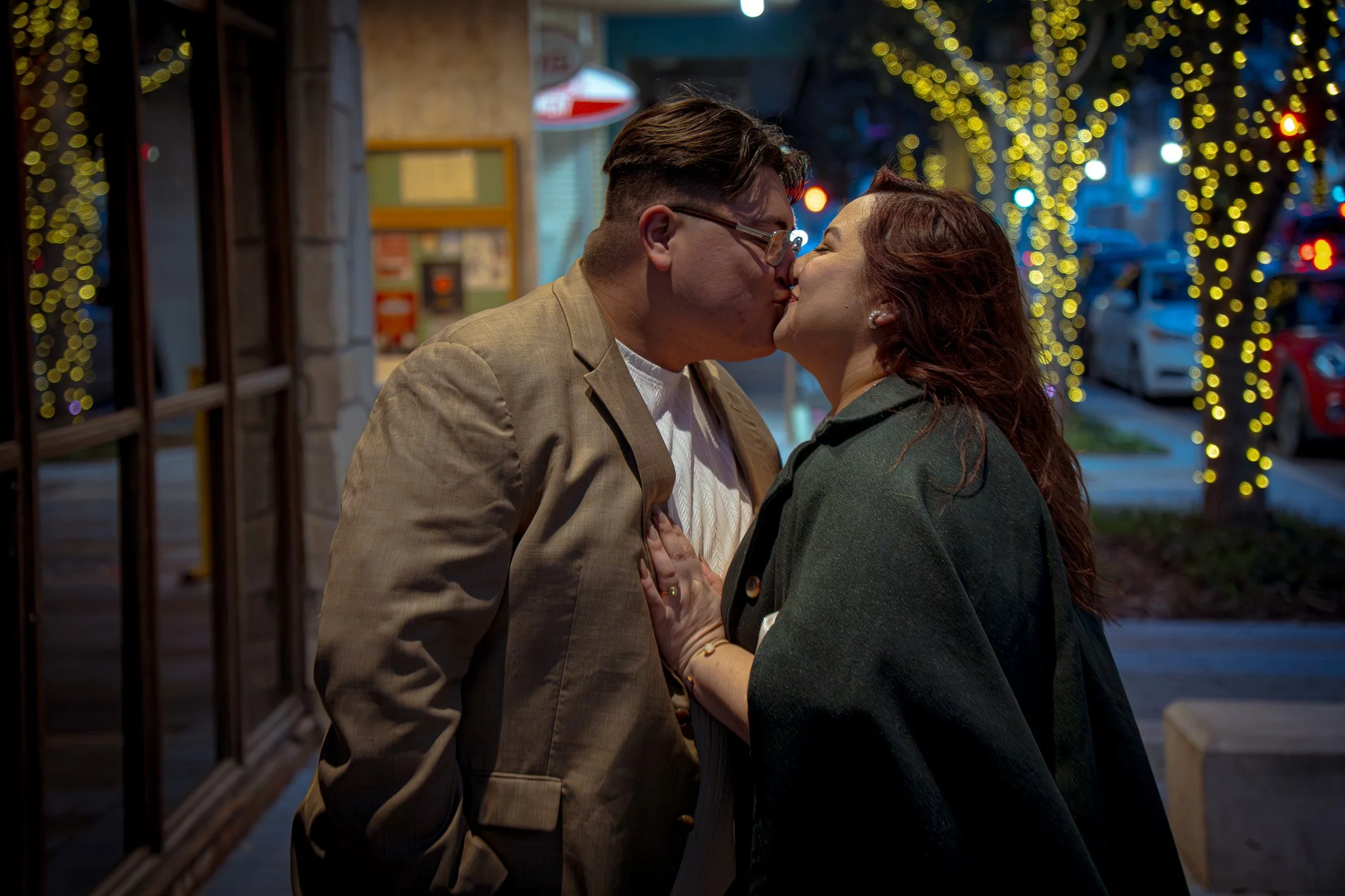 A couple sharing a kiss on a city sidewalk at night, with Christmas lights on trees in the background.
