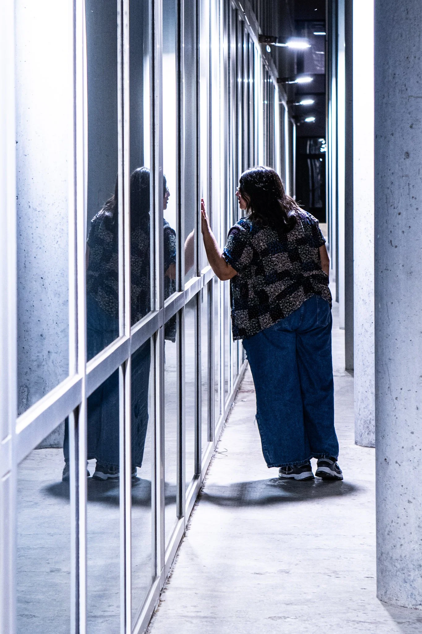 A woman with dark hair wearing a patterned shirt and loose jeans standing in front of a glass wall, touching it with her hand, with her reflection visible in the glass.