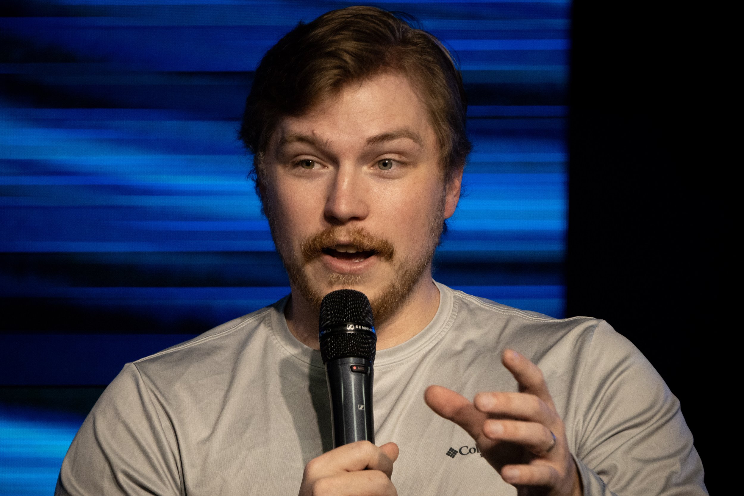 A young man with brown hair and a beard holding a microphone and speaking in front of a blue striped background.