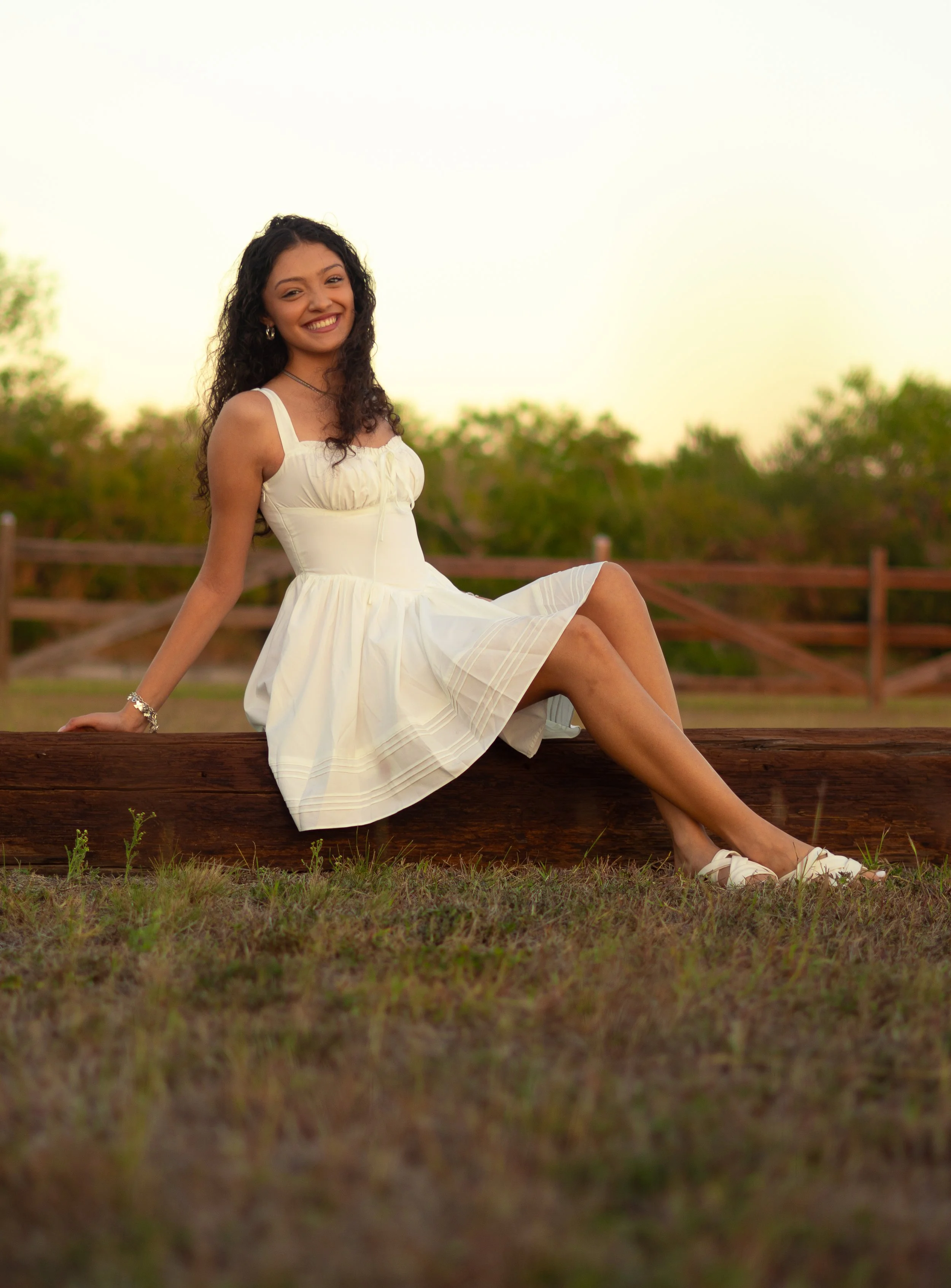 A young woman with curly dark hair sitting on a wooden fence outdoors at sunset, smiling at the camera, wearing a white sundress and white shoes.