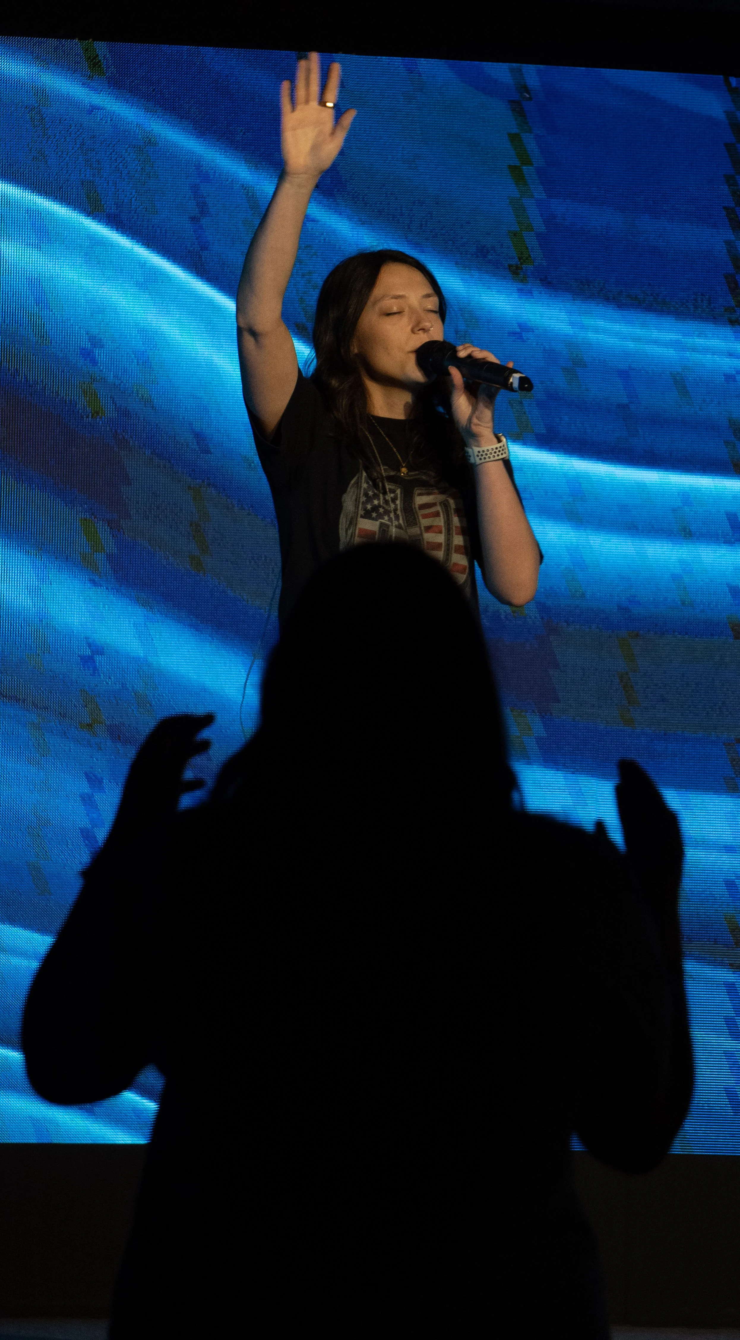 A woman singing on stage with her eyes closed, holding a microphone in her right hand, and raising her left hand. There is a person in the foreground with hands raised, silhouetted against a large digital screen with blue and green graphics.