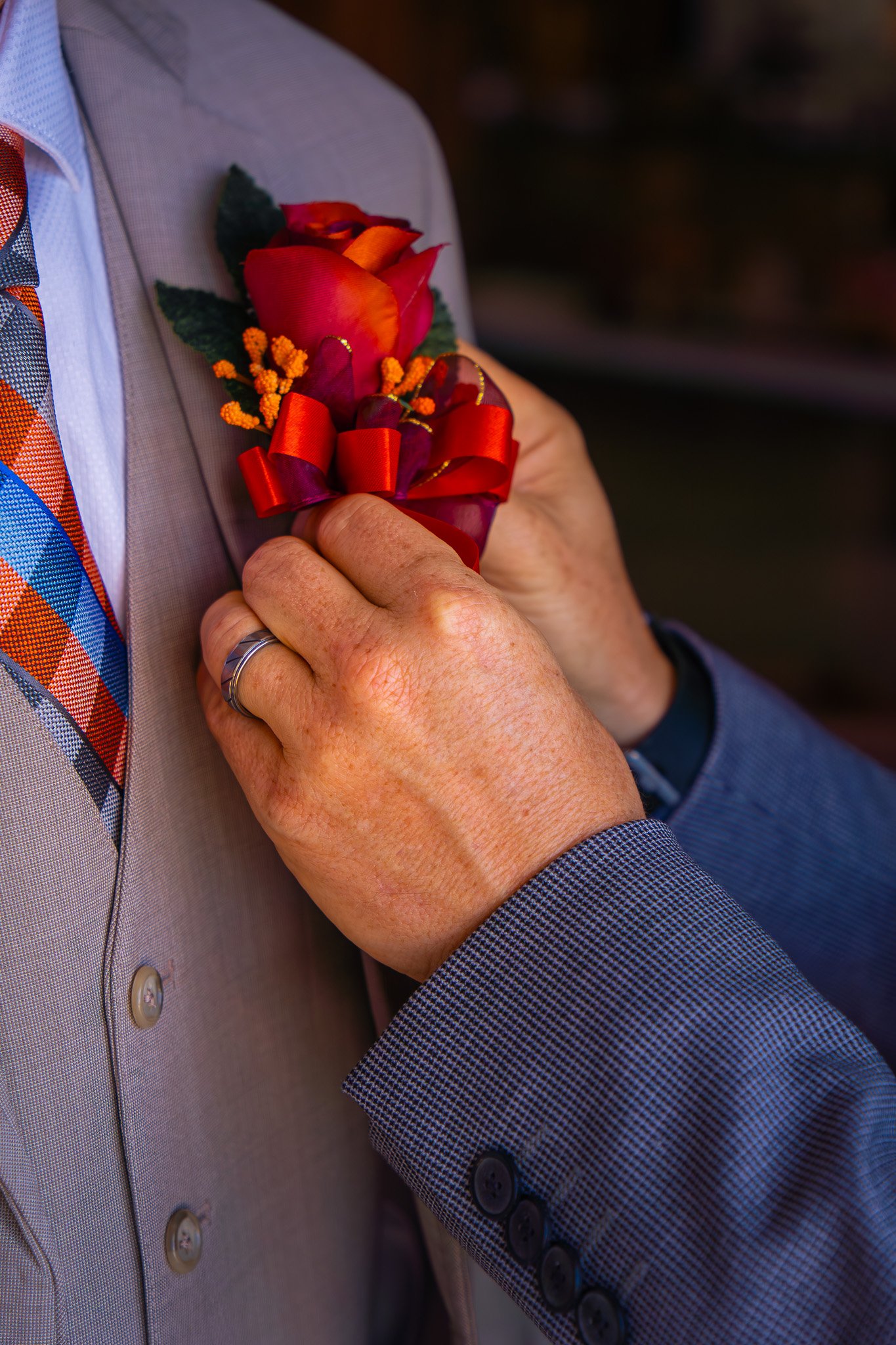 Close-up of a man in a suit pinning on a red and orange boutonniere with a red ribbon, with a checkered shirt and patterned tie, during a formal or wedding event.